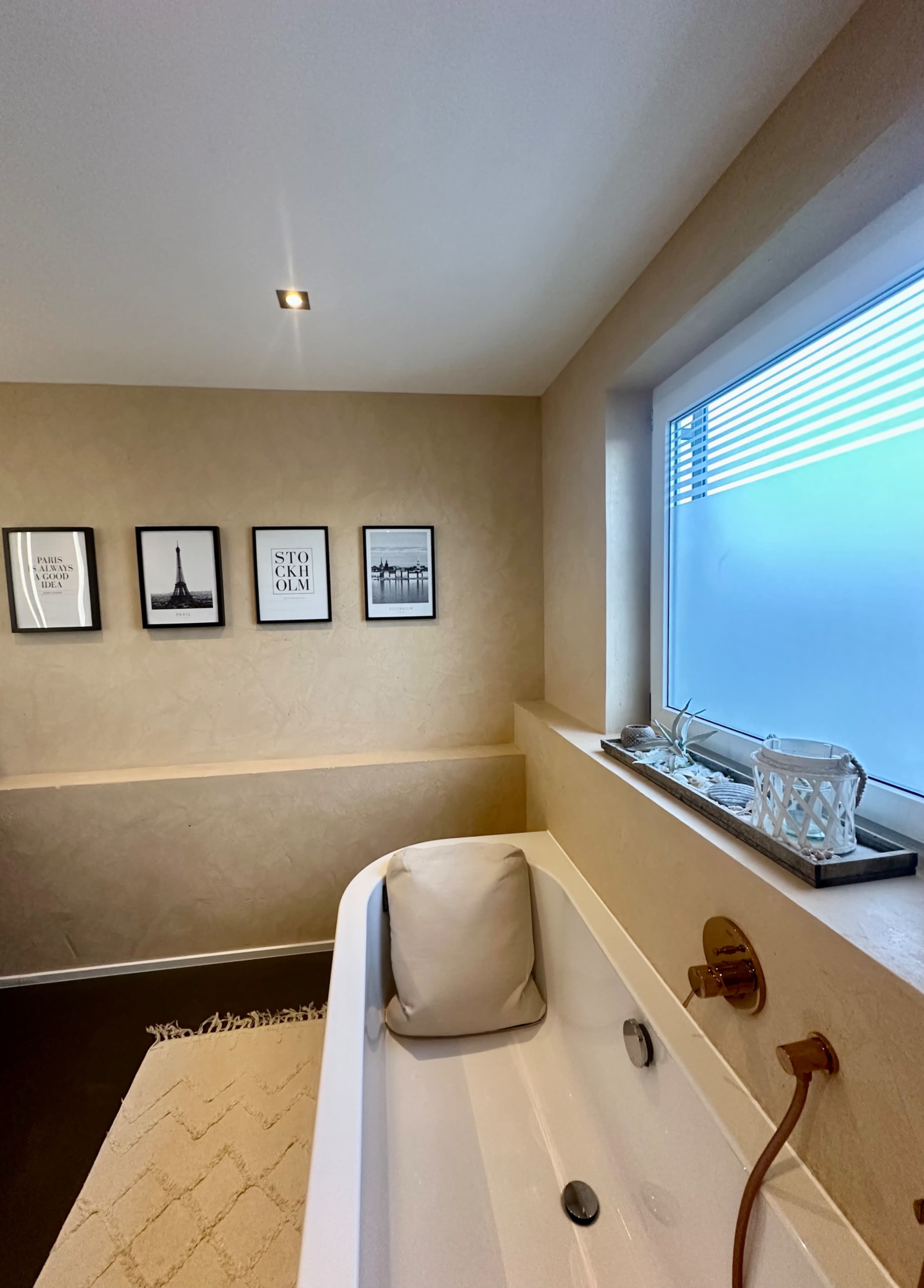 The image shows a modern bathroom with a bathtub, a window with horizontal blinds, and framed black-and-white photographs on the wall.