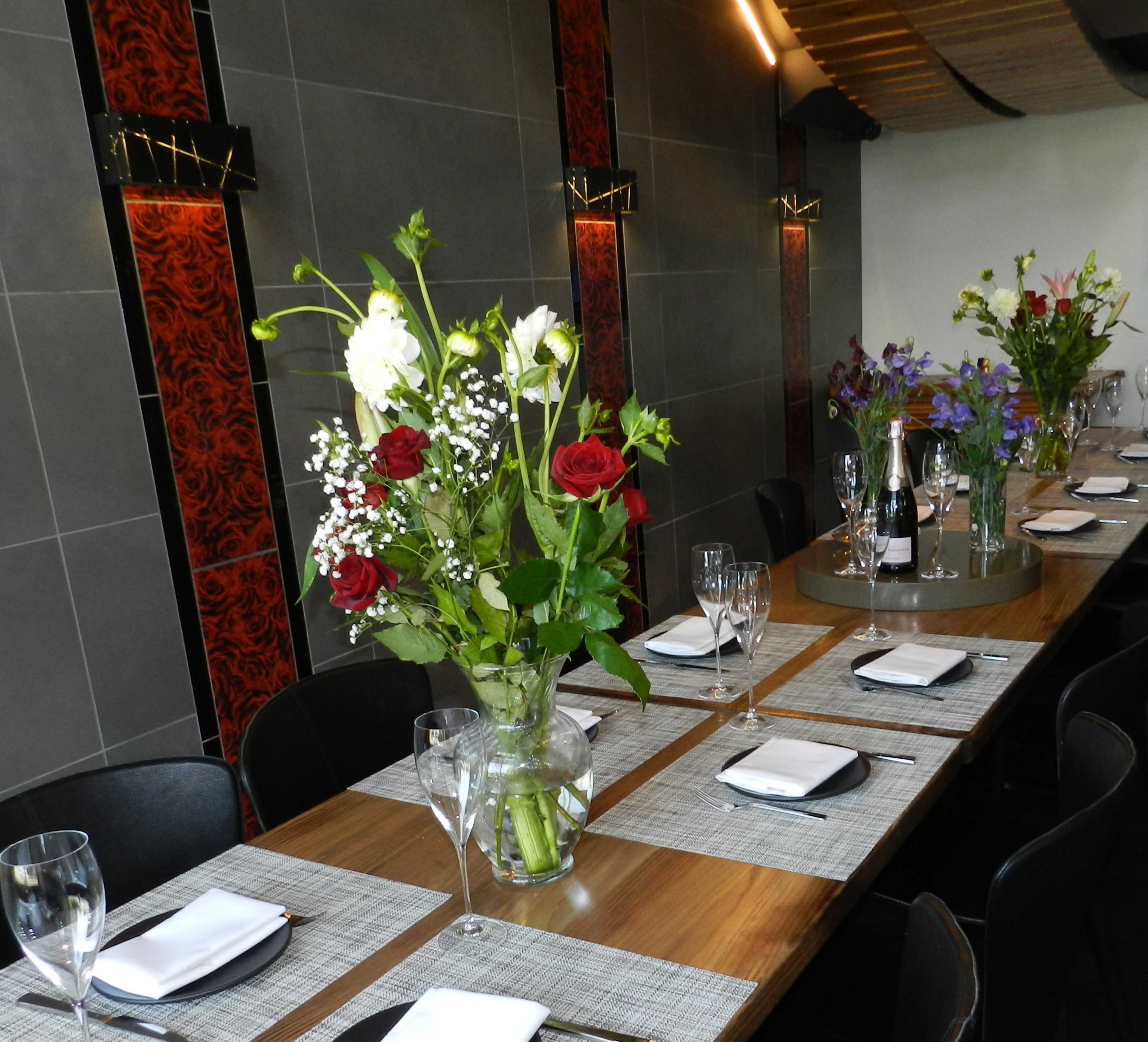 A long dining table is set with silverware and glasses, topped with a large vase of flowers in a modern restaurant interior.