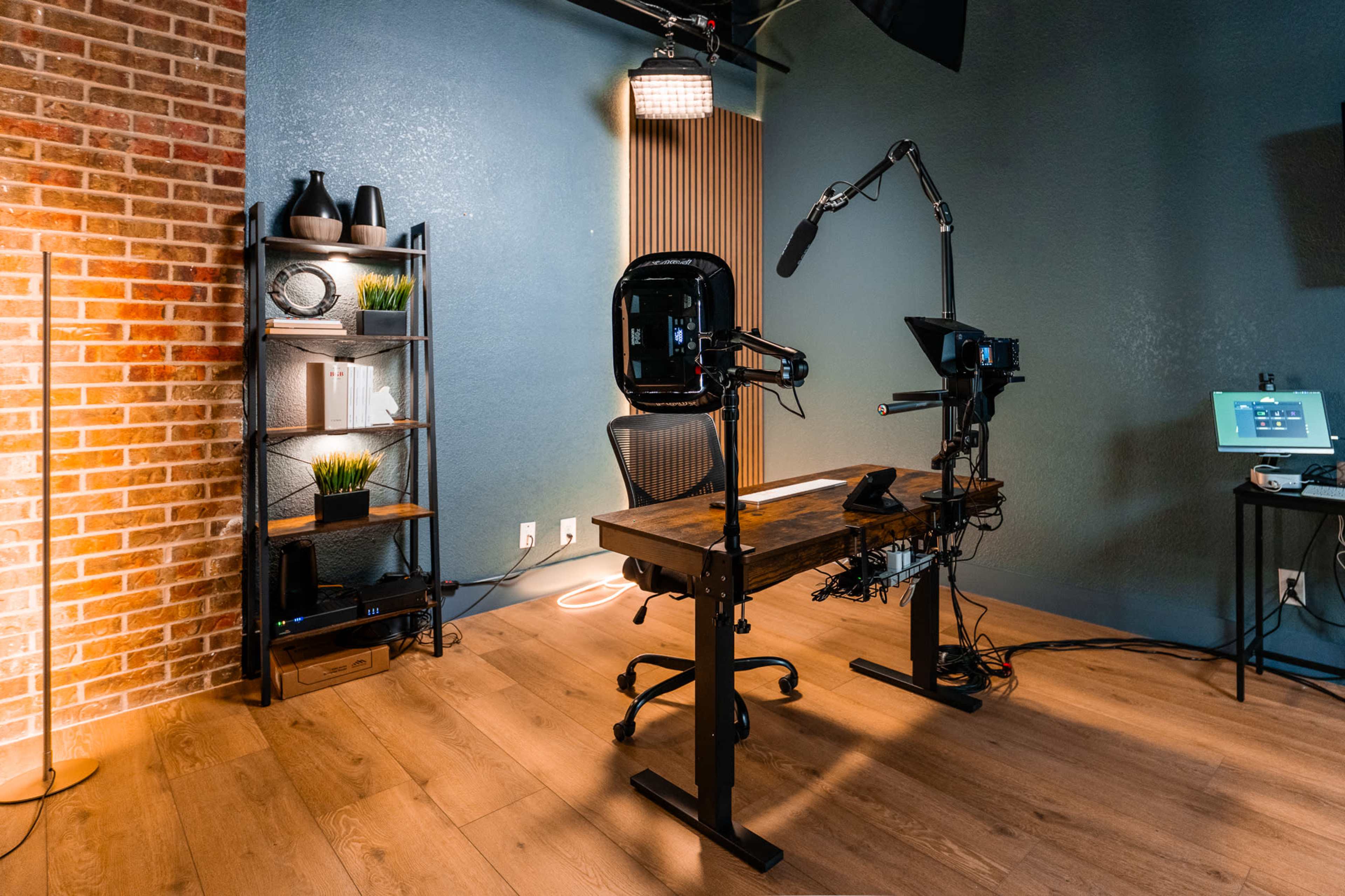 The image shows a modern studio setup with a wooden desk, a camera on a tripod, and a shelf displaying decorative items against a blue wall.