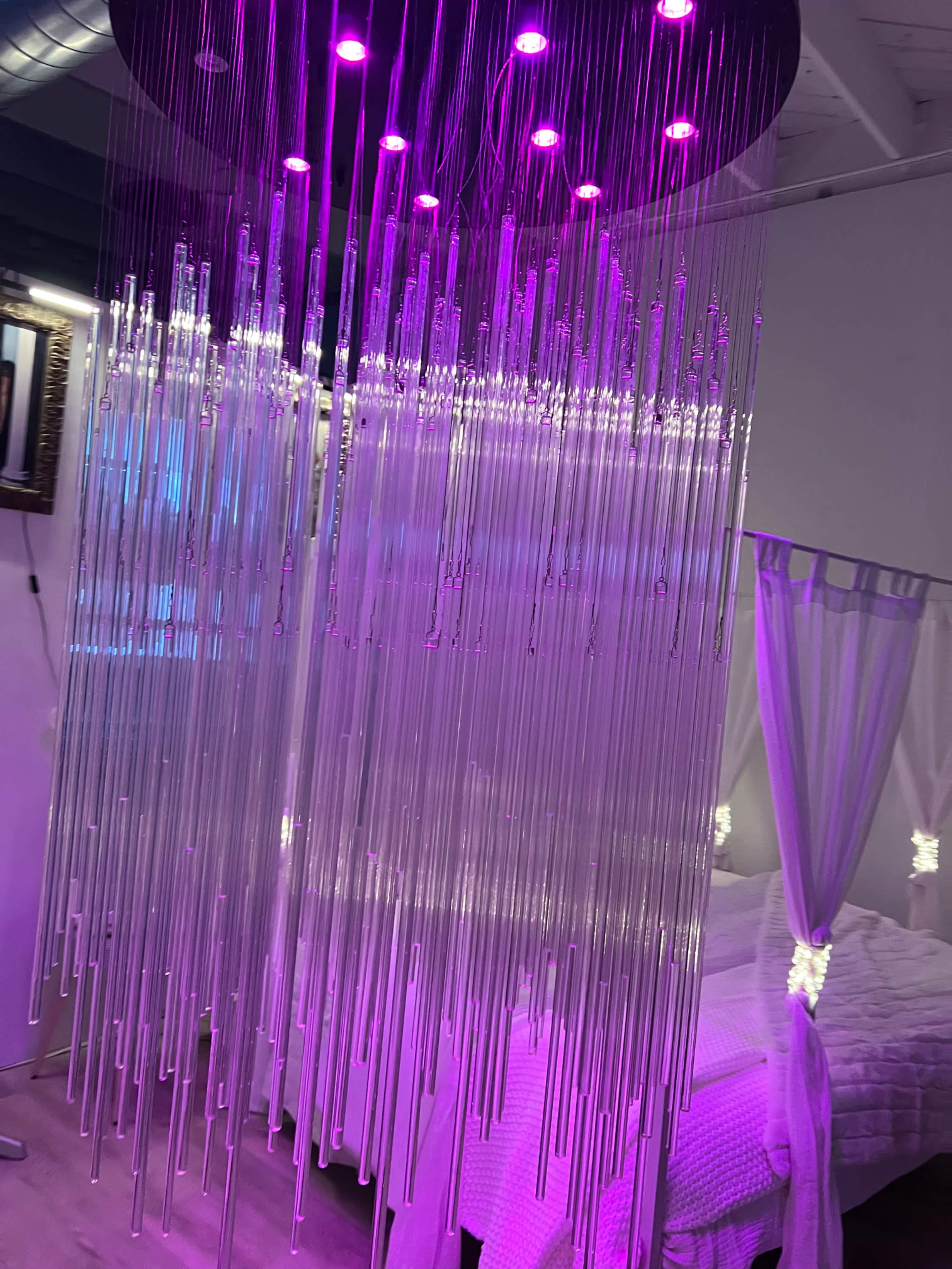 The image shows a modern bedroom with a translucent, hanging installation featuring vertical strands illuminated by purple lights.