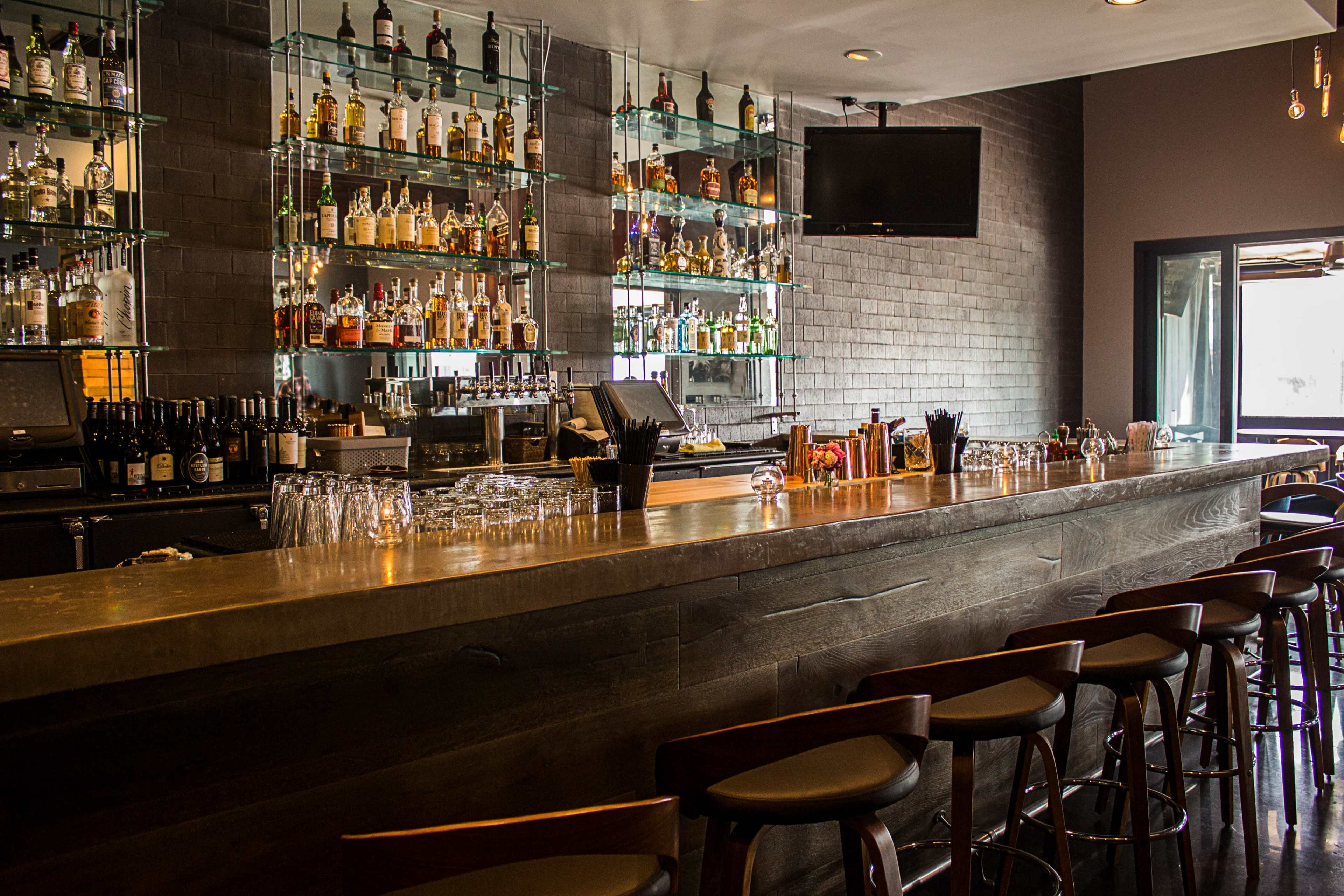 A modern bar with a long countertop, shelves displaying numerous bottles of liquor, and a television mounted on the wall.