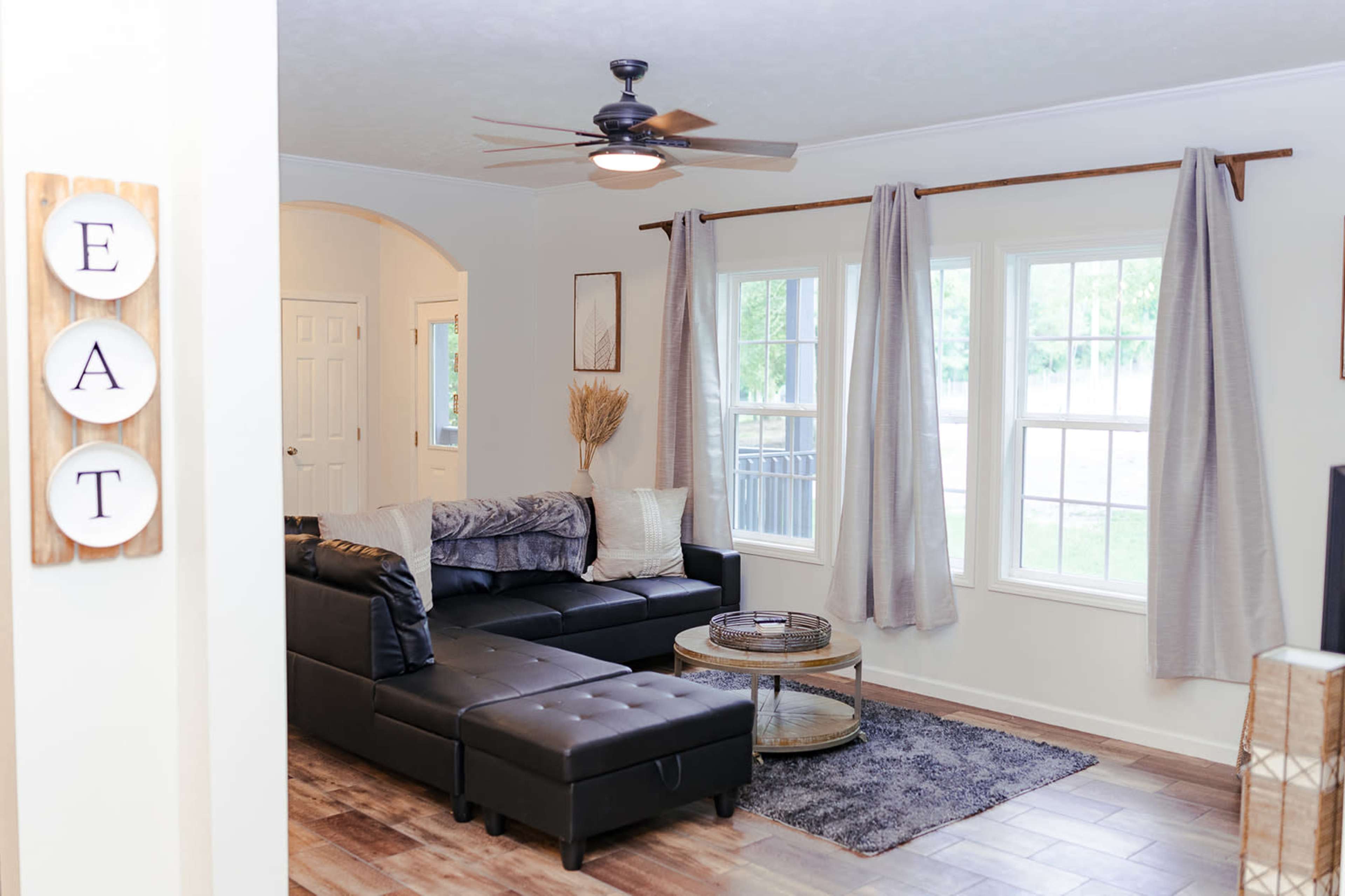 A cozy living room featuring a black sectional sofa, a round coffee table, and large windows with gray curtains.