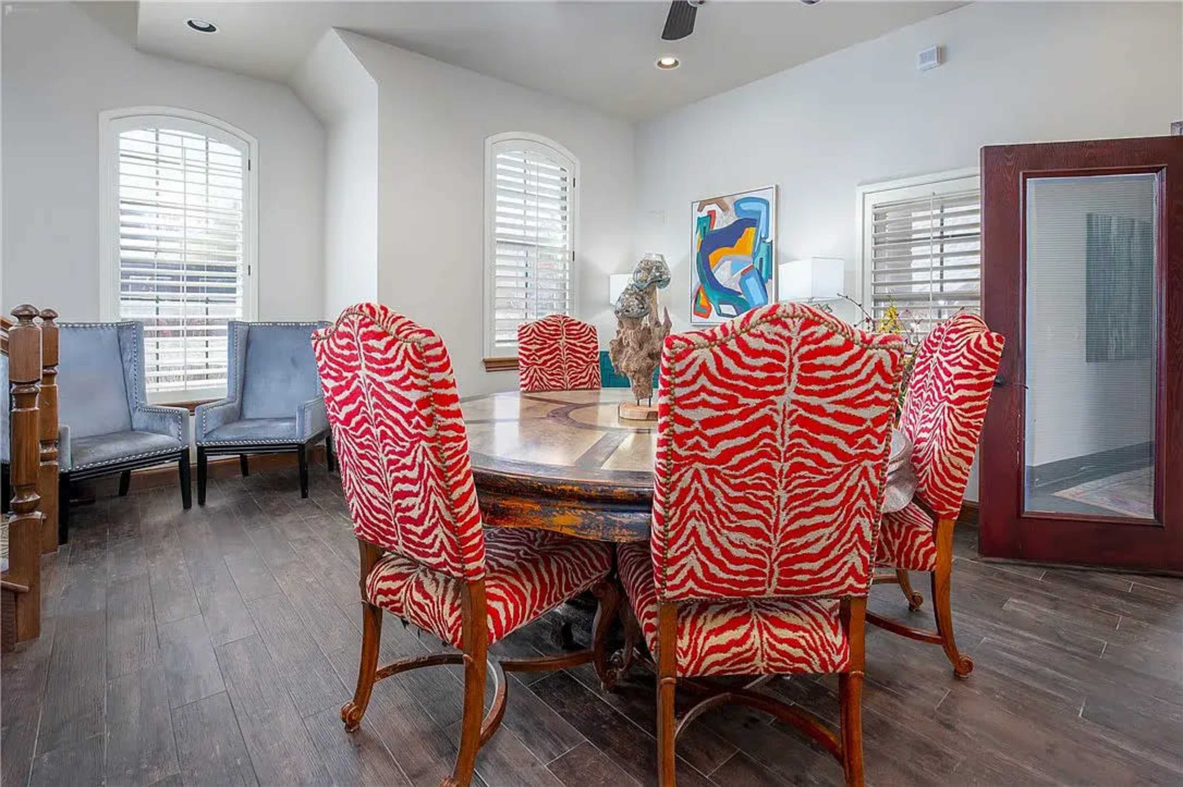 A round dining table surrounded by six chairs with a bold red and white zebra pattern, situated in a well-lit room with large windows and decorative art.