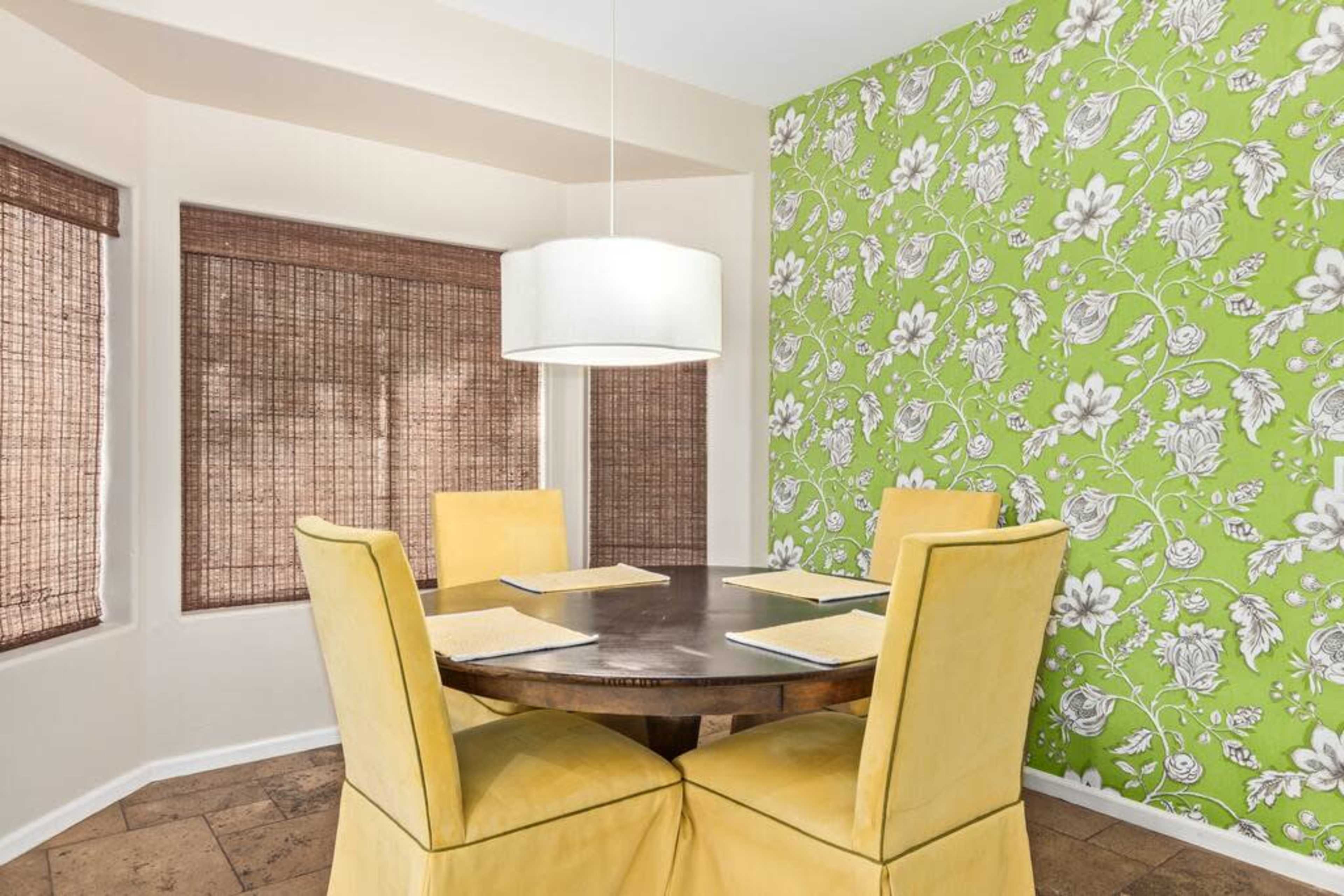 A round dining table with four yellow chairs is set in a room with a green floral-patterned accent wall and bamboo window shades.