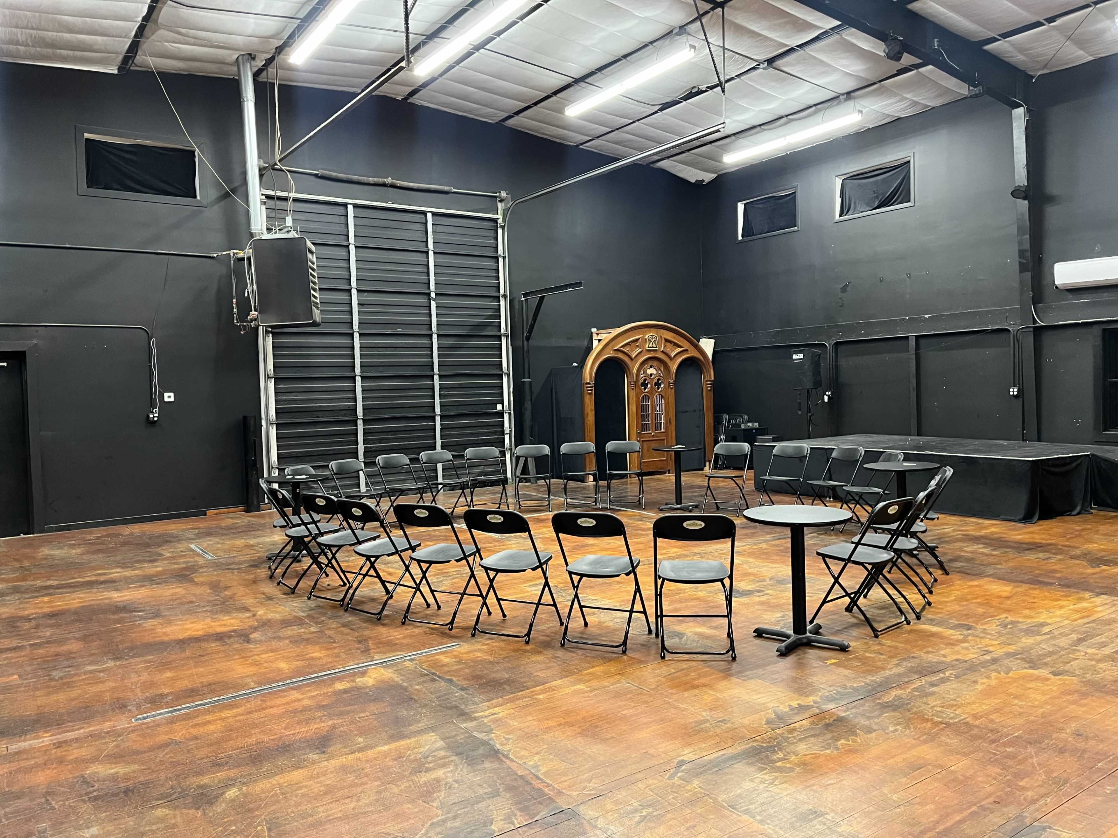 The image shows an empty rehearsal space with a circular arrangement of chairs around two small tables and a decorative wooden structure in the background.