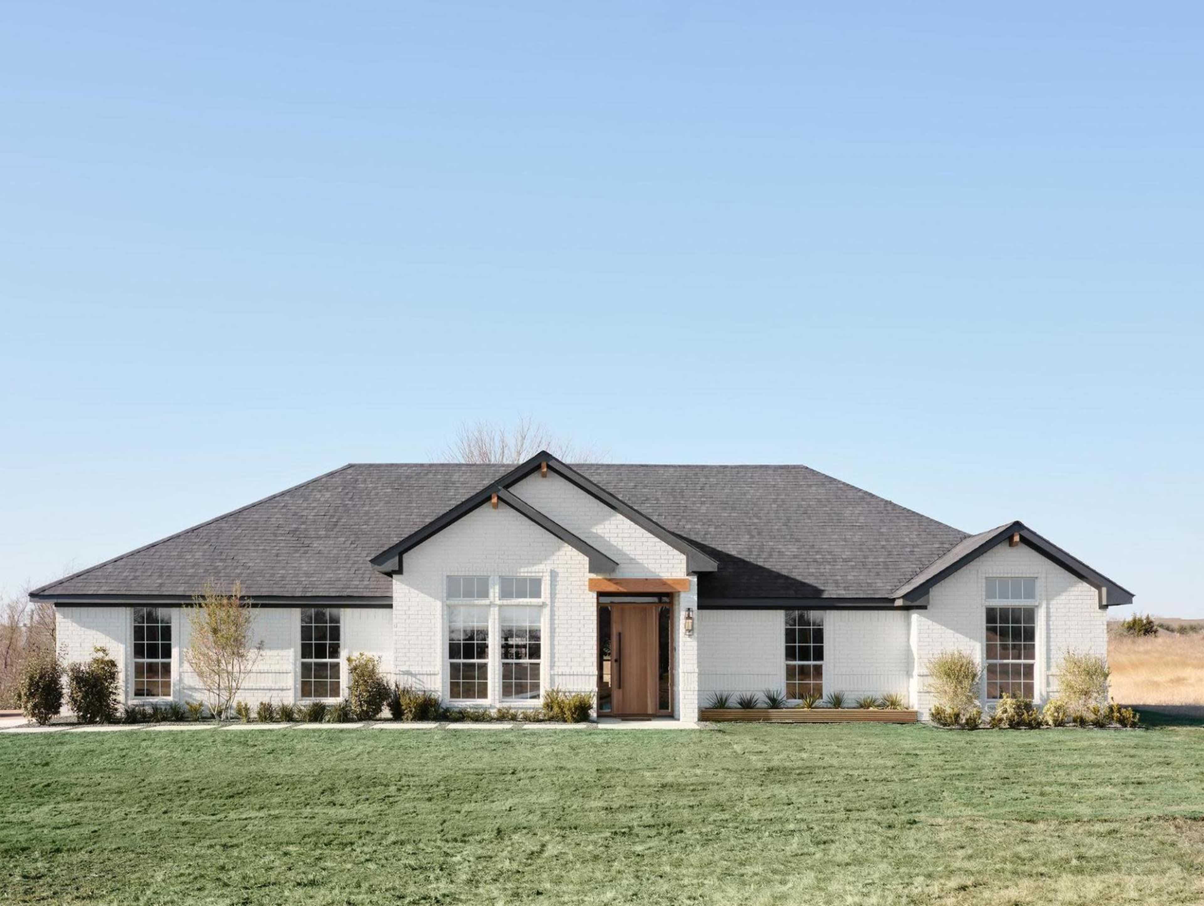 A modern single-story house with a white brick exterior and a dark roof, situated on a green lawn under a clear blue sky.