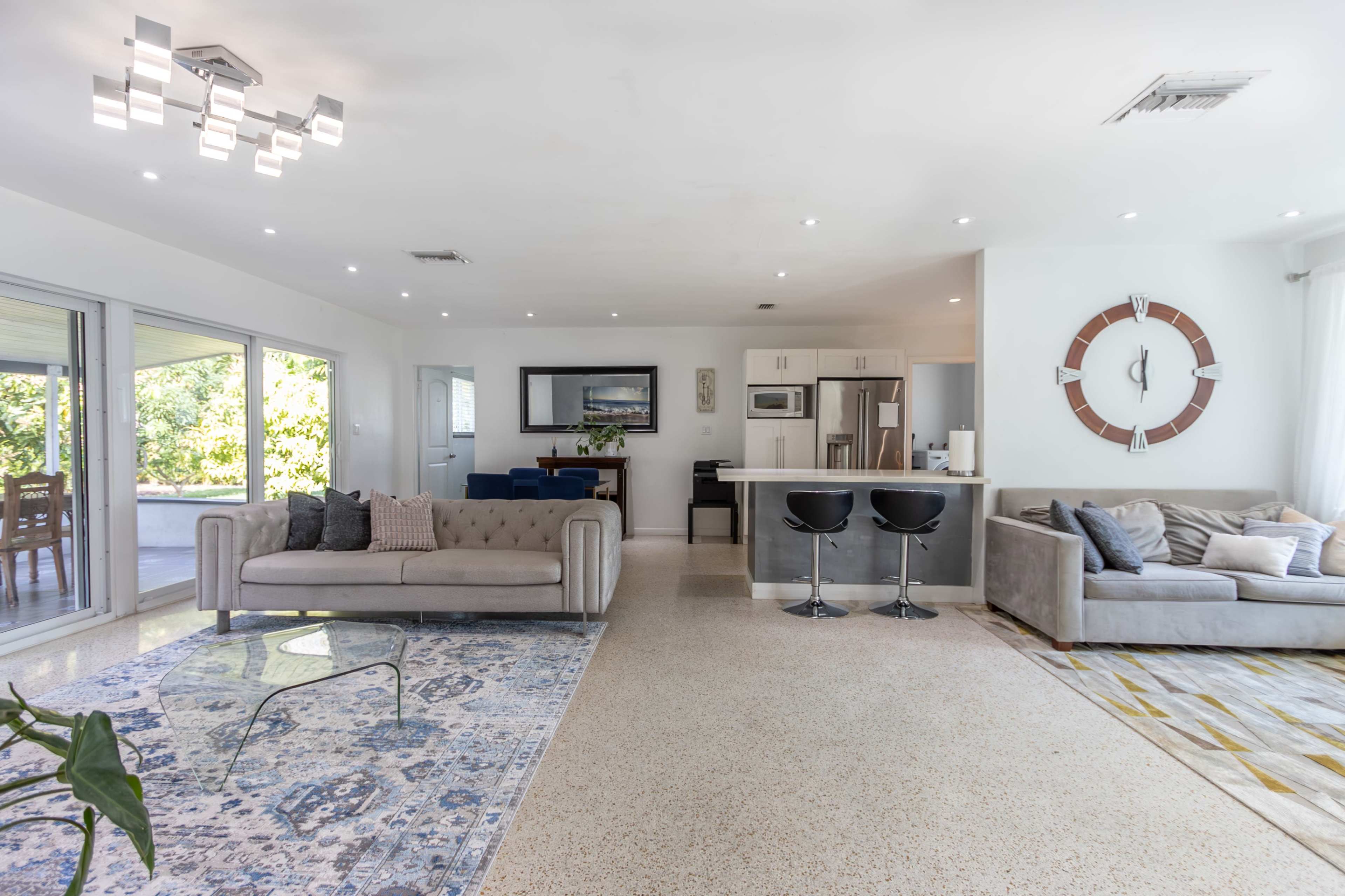 A modern living space featuring a light-colored sectional sofa, a gray sofa, a patterned rug, and a sleek kitchen area with bar stools.
