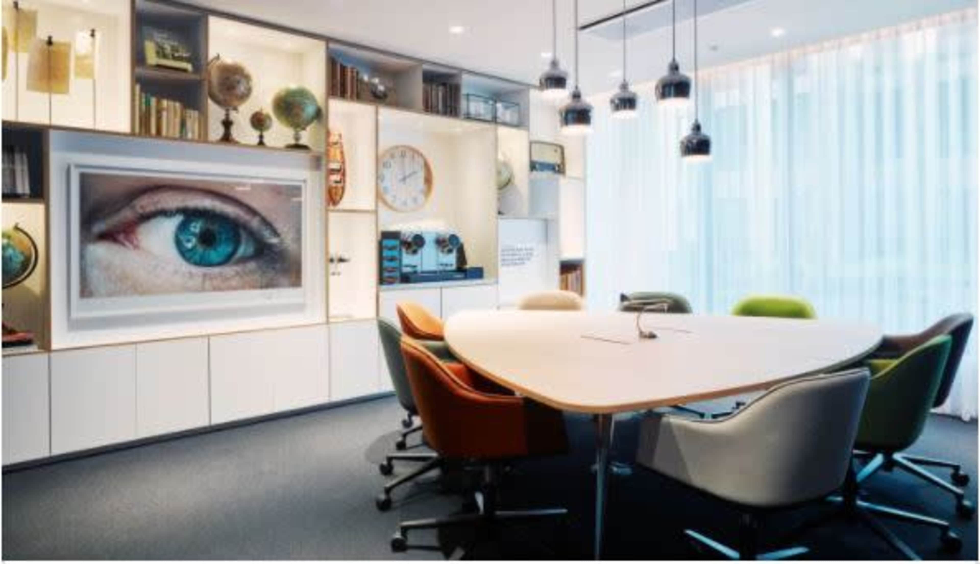 A modern conference room with a large table surrounded by colorful chairs, and a display featuring an eye on the wall among shelves filled with decorative items and a clock.