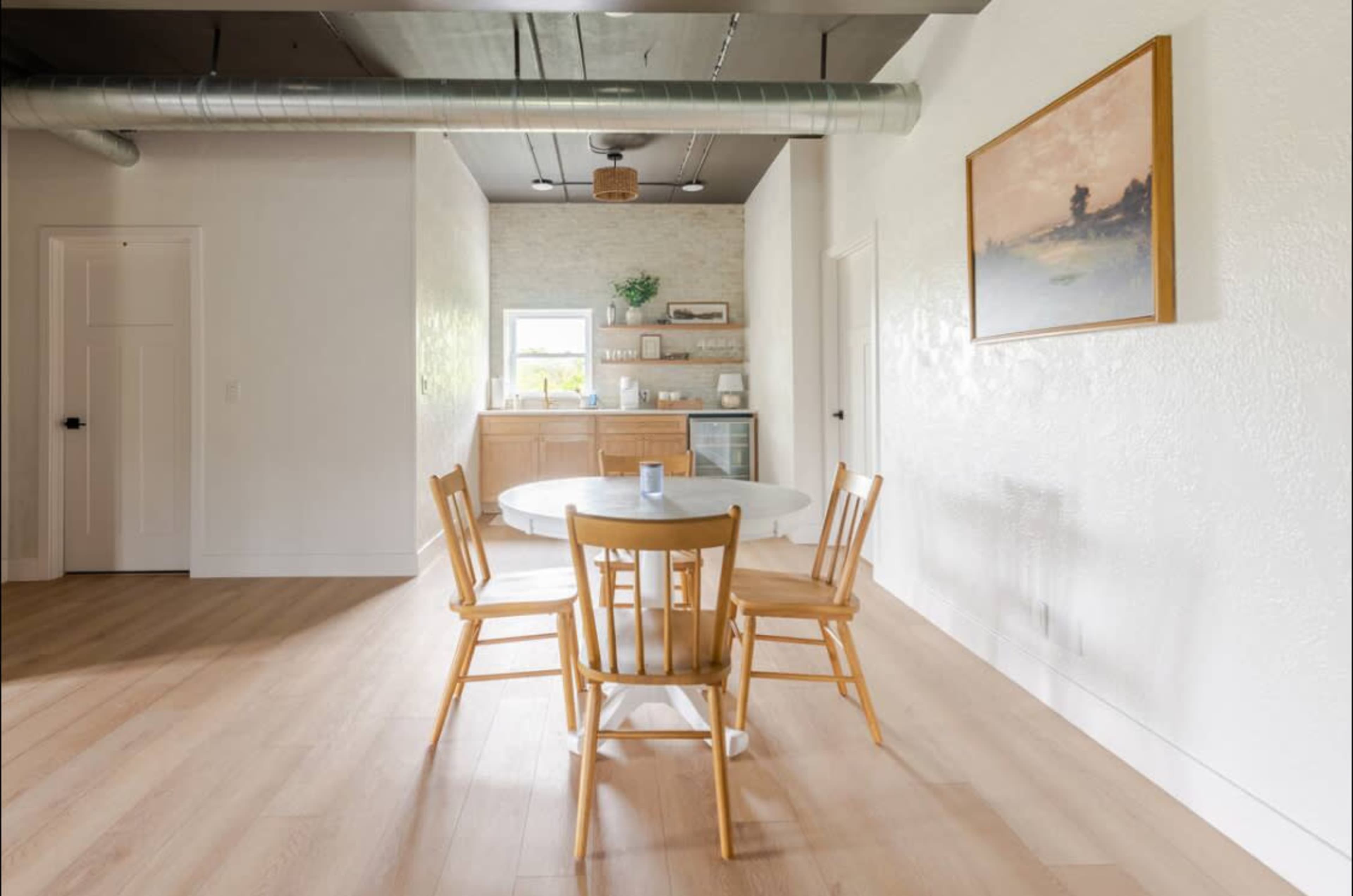 A round table with four wooden chairs is positioned in a bright dining area featuring a kitchen in the background.