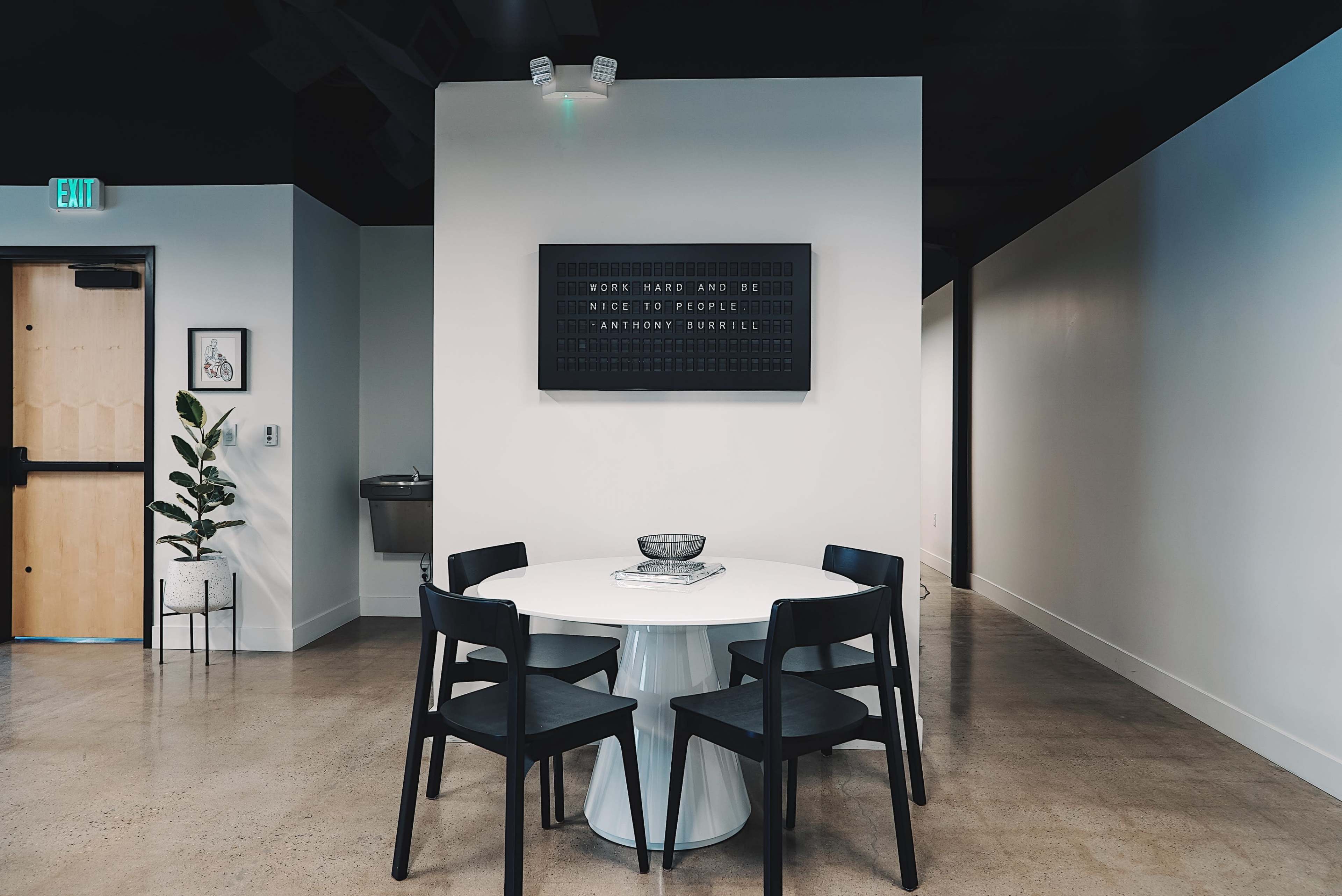 A round white table with four black chairs is centered on a light-colored floor beneath a wall-mounted board featuring text.