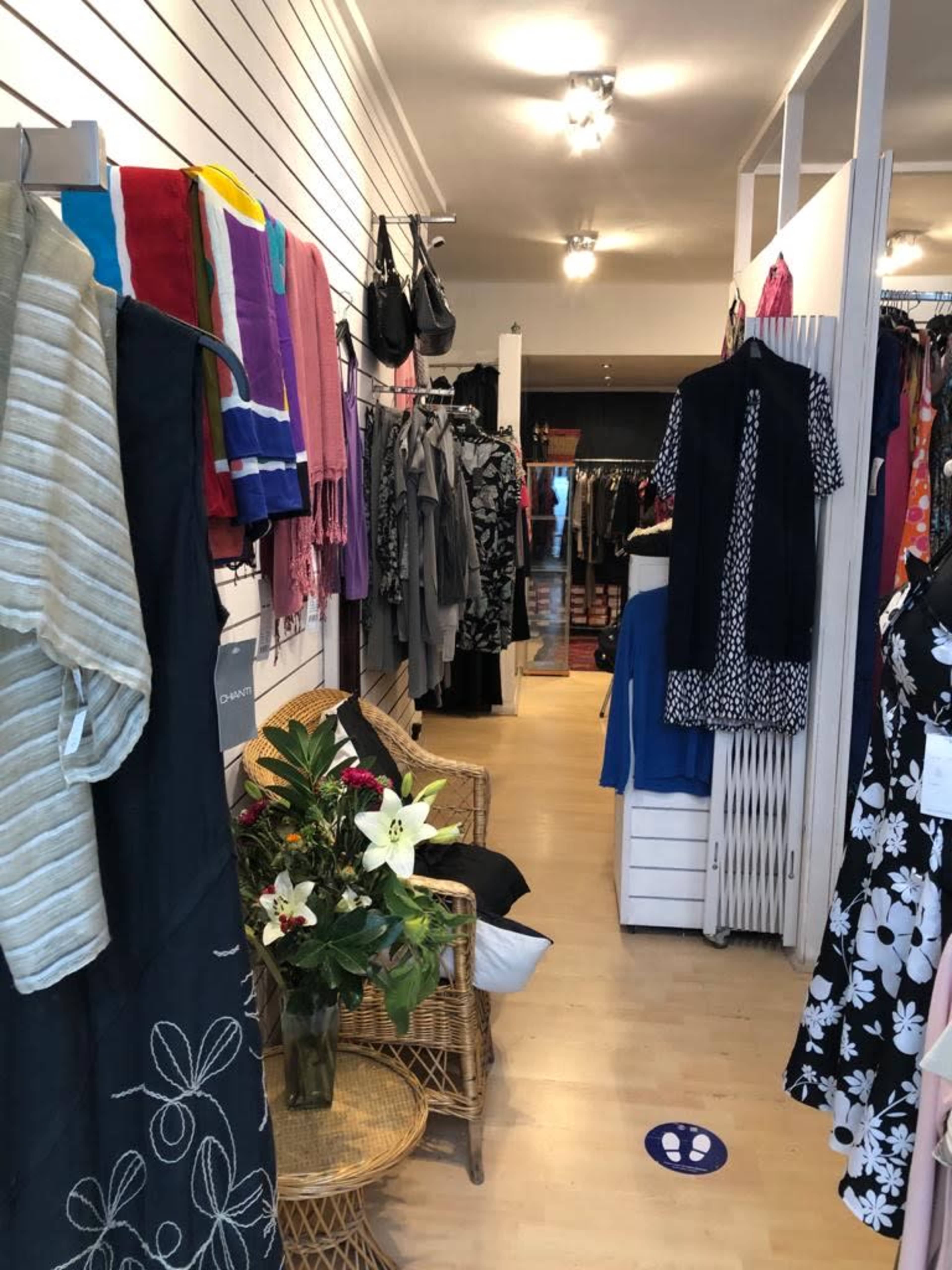 The image shows the interior of a clothing store with racks of apparel lining the walls and a seating area featuring a plant.