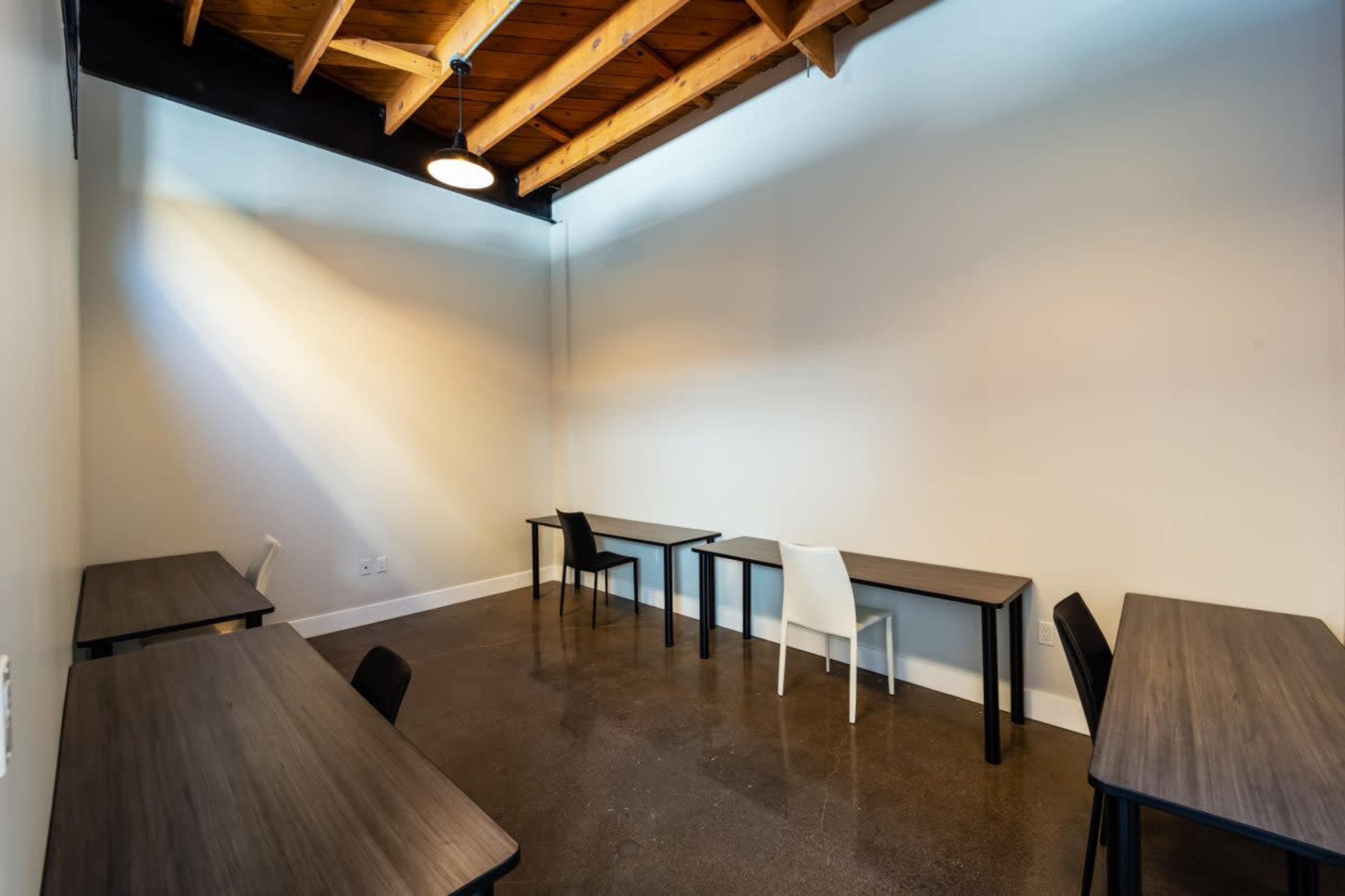 The image shows a minimalist room with several wooden tables and chairs, featuring a high ceiling with exposed beams and polished concrete flooring.