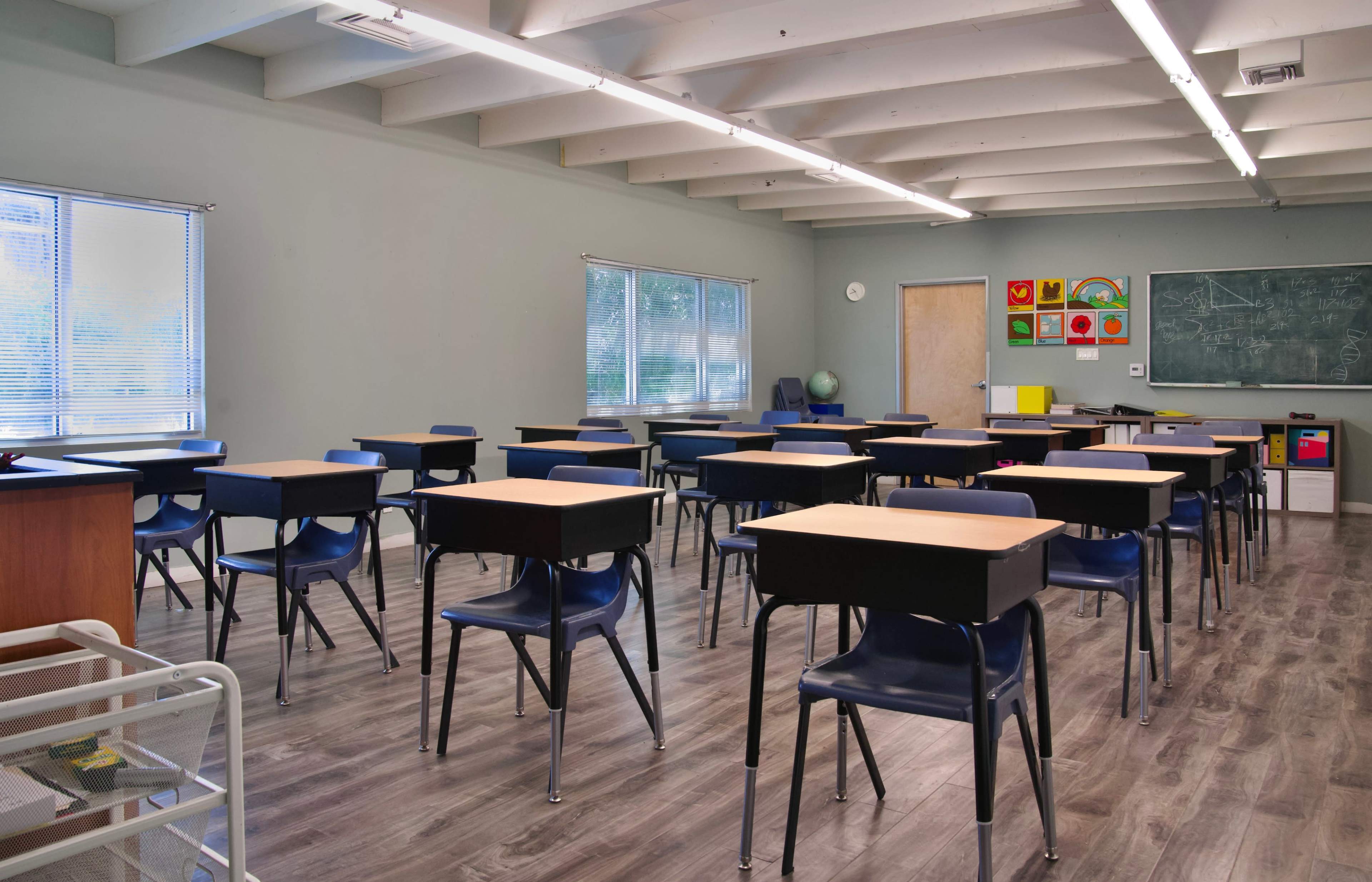 The classroom features rows of empty black desks and blue chairs, with a chalkboard and educational posters on the walls.