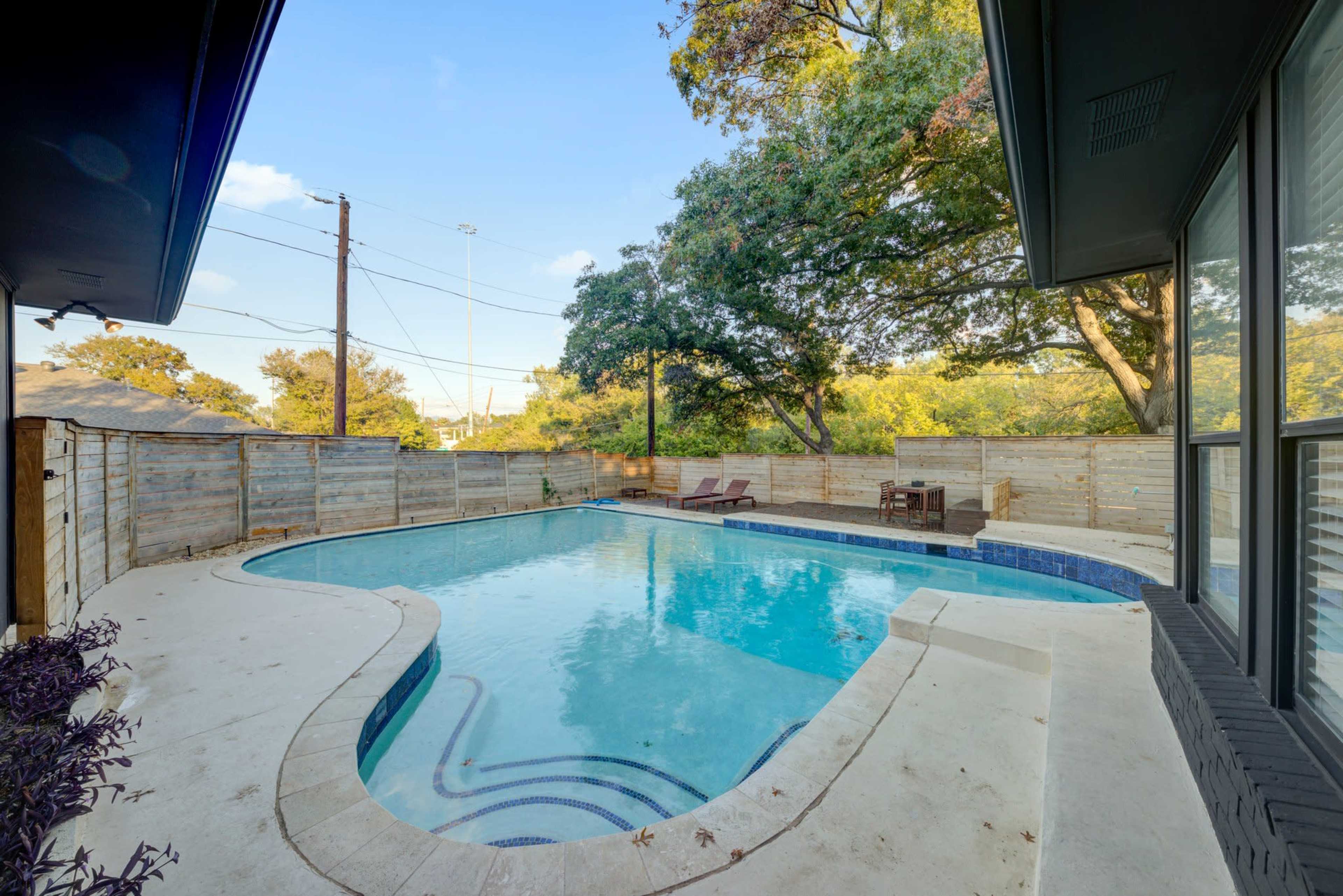 The image shows a clear swimming pool surrounded by a wooden fence and a patio area with seating.