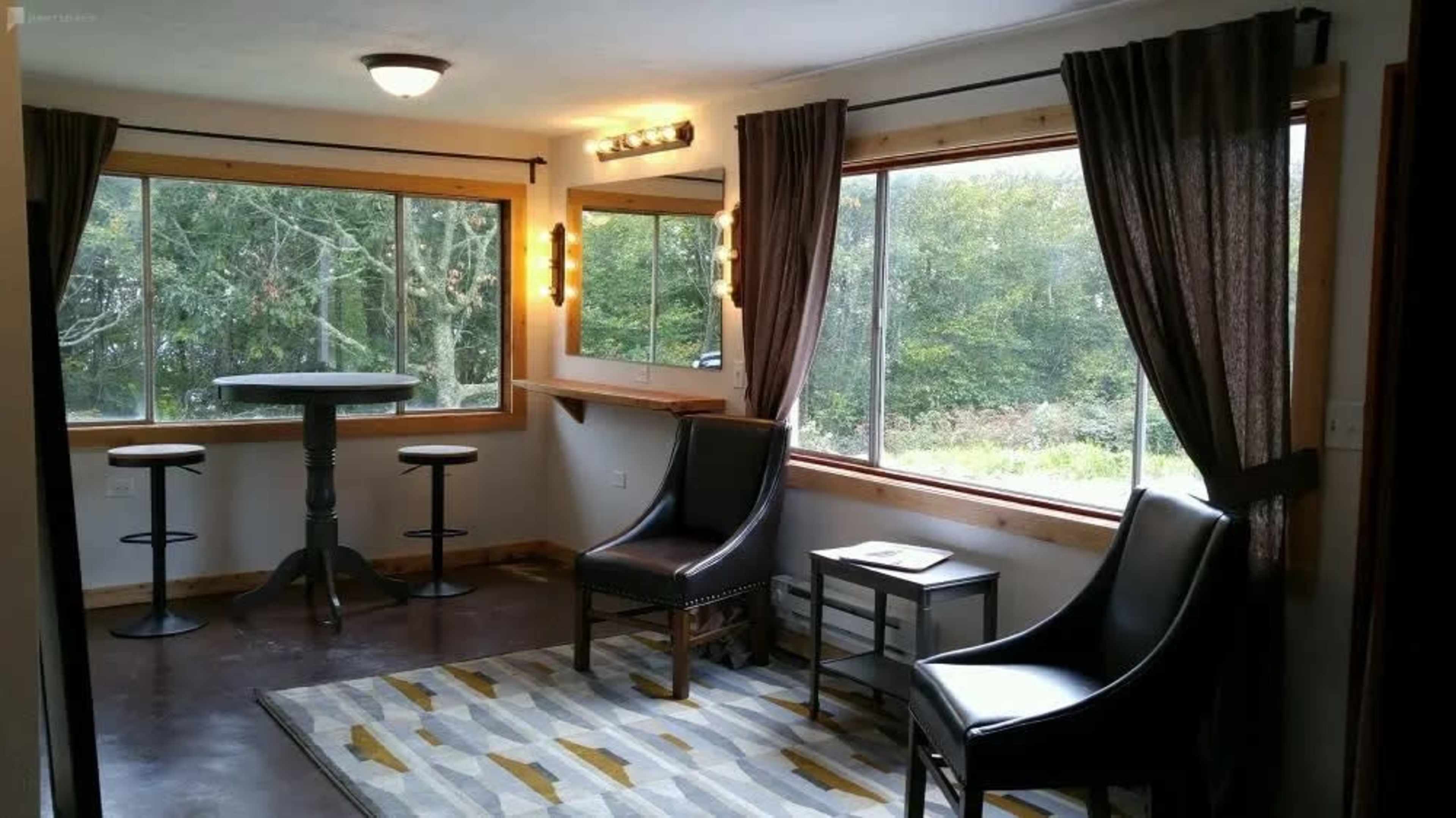 A cozy room with two black chairs, a circular table with three stools, and large windows overlooking a wooded area.