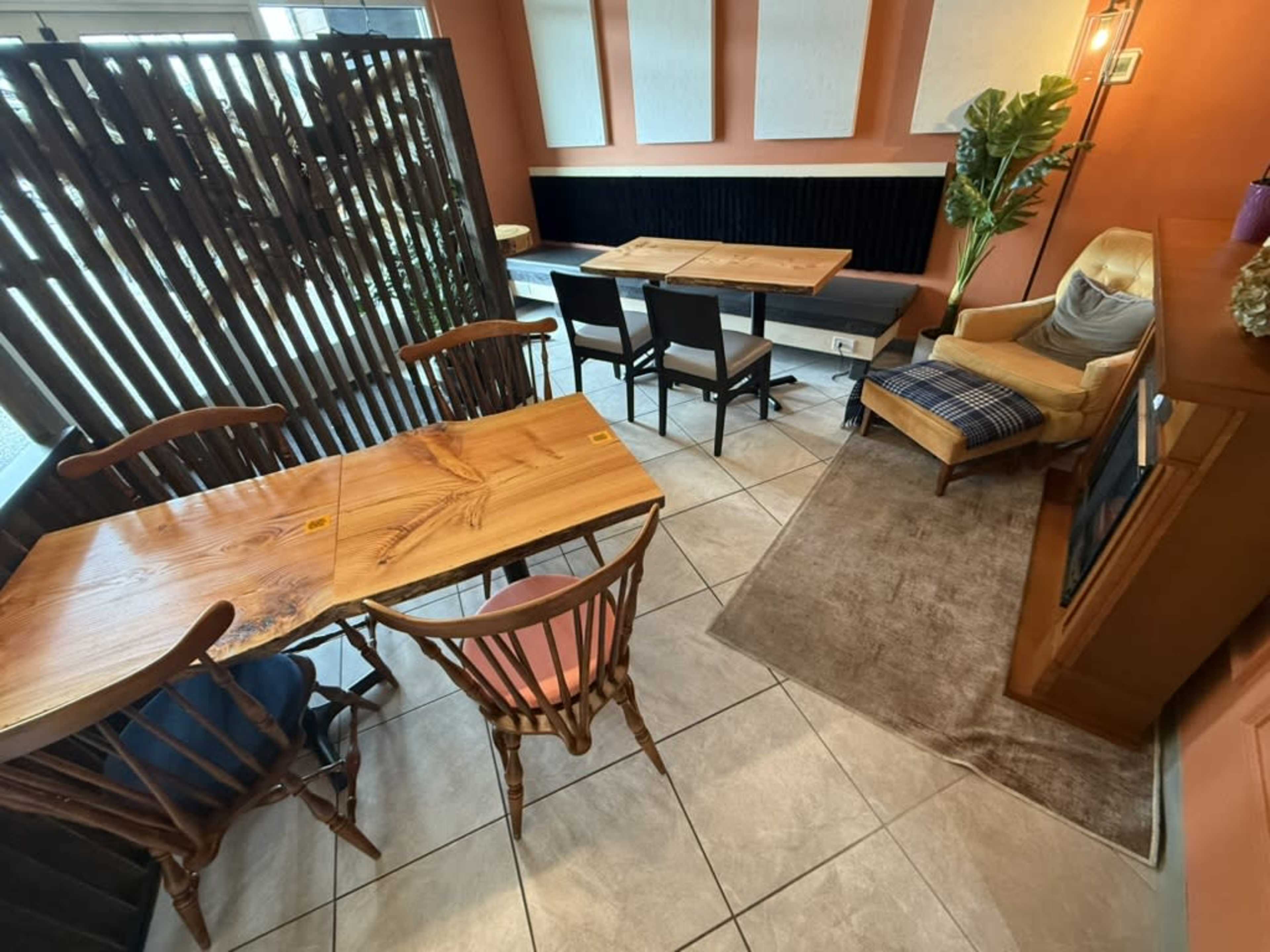 The image shows a cozy café interior with wooden furniture, including tables and chairs, a small rug, and a lounge chair in a corner.
