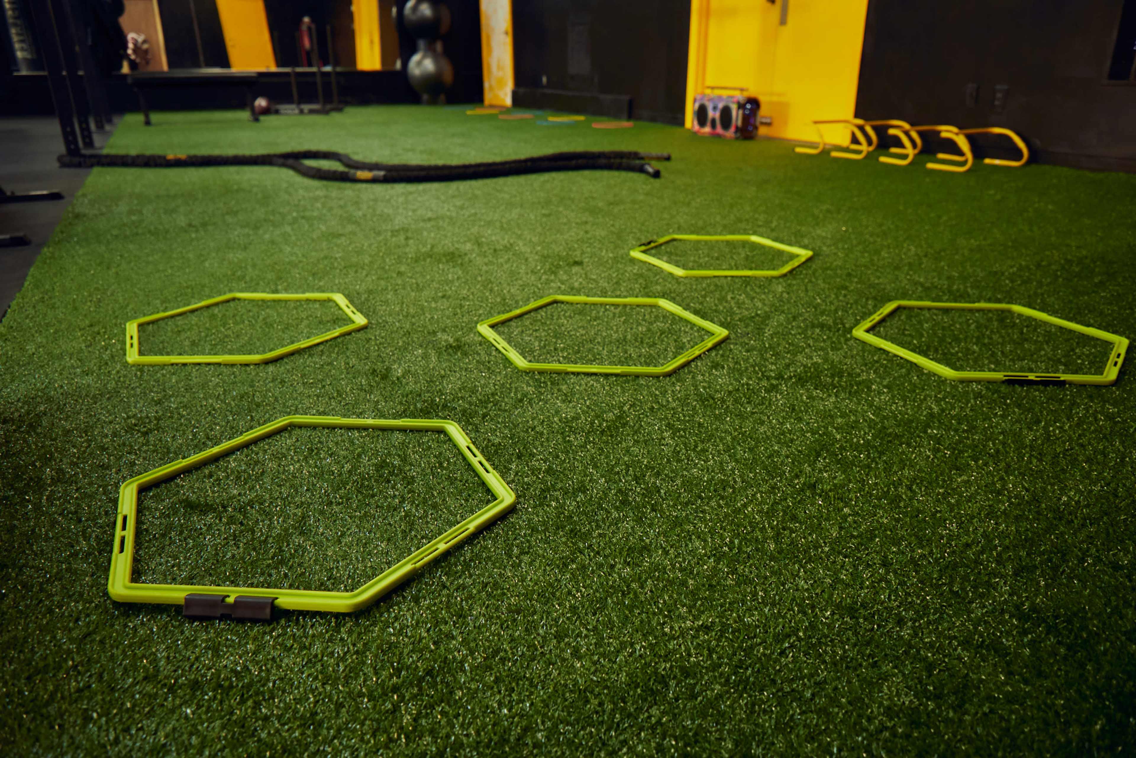 Five green hexagonal training cones are arranged on a synthetic turf surface in a gym setting.