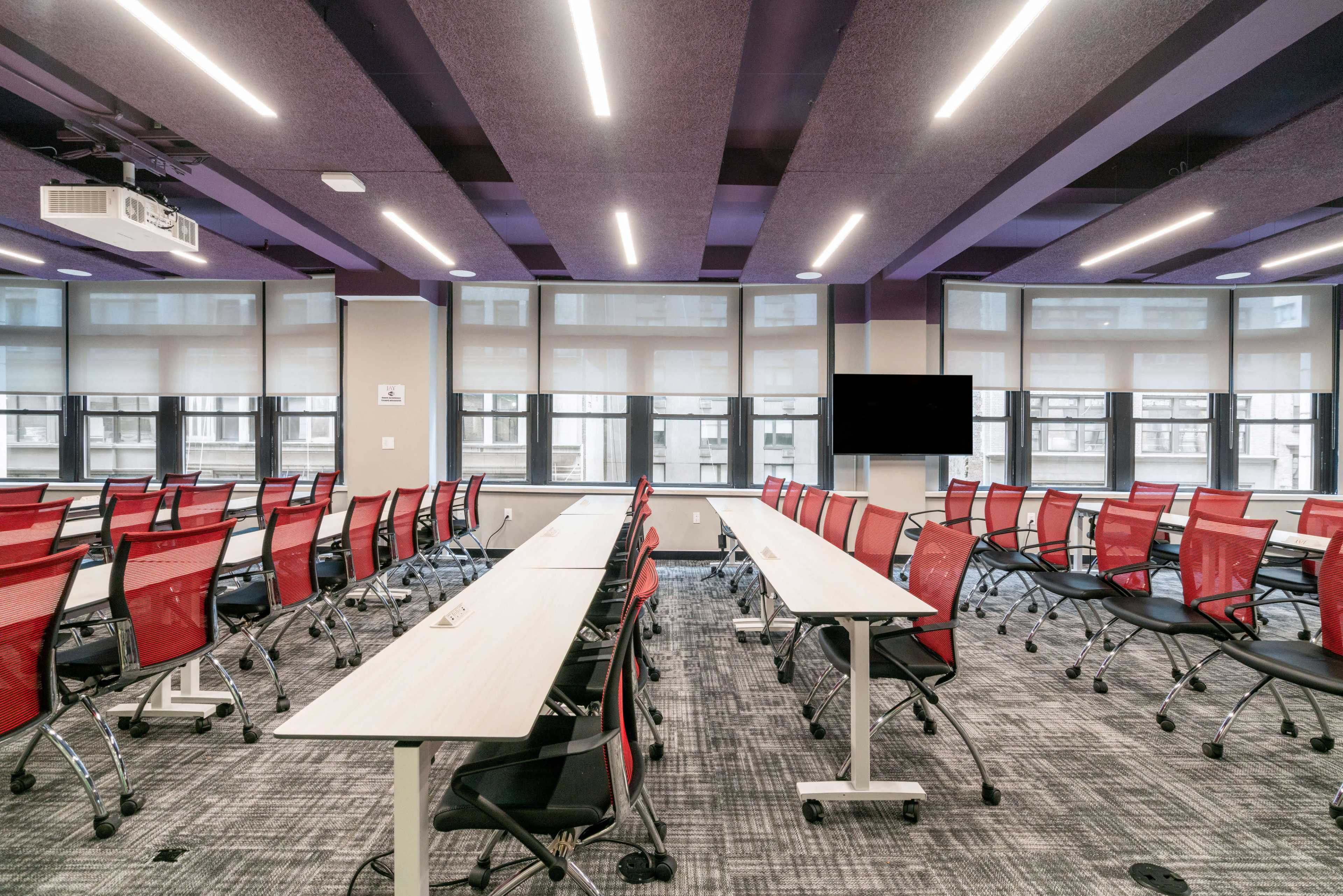 A modern classroom features rows of red and black chairs with adjustable white tables, illuminated by overhead lights and large windows.