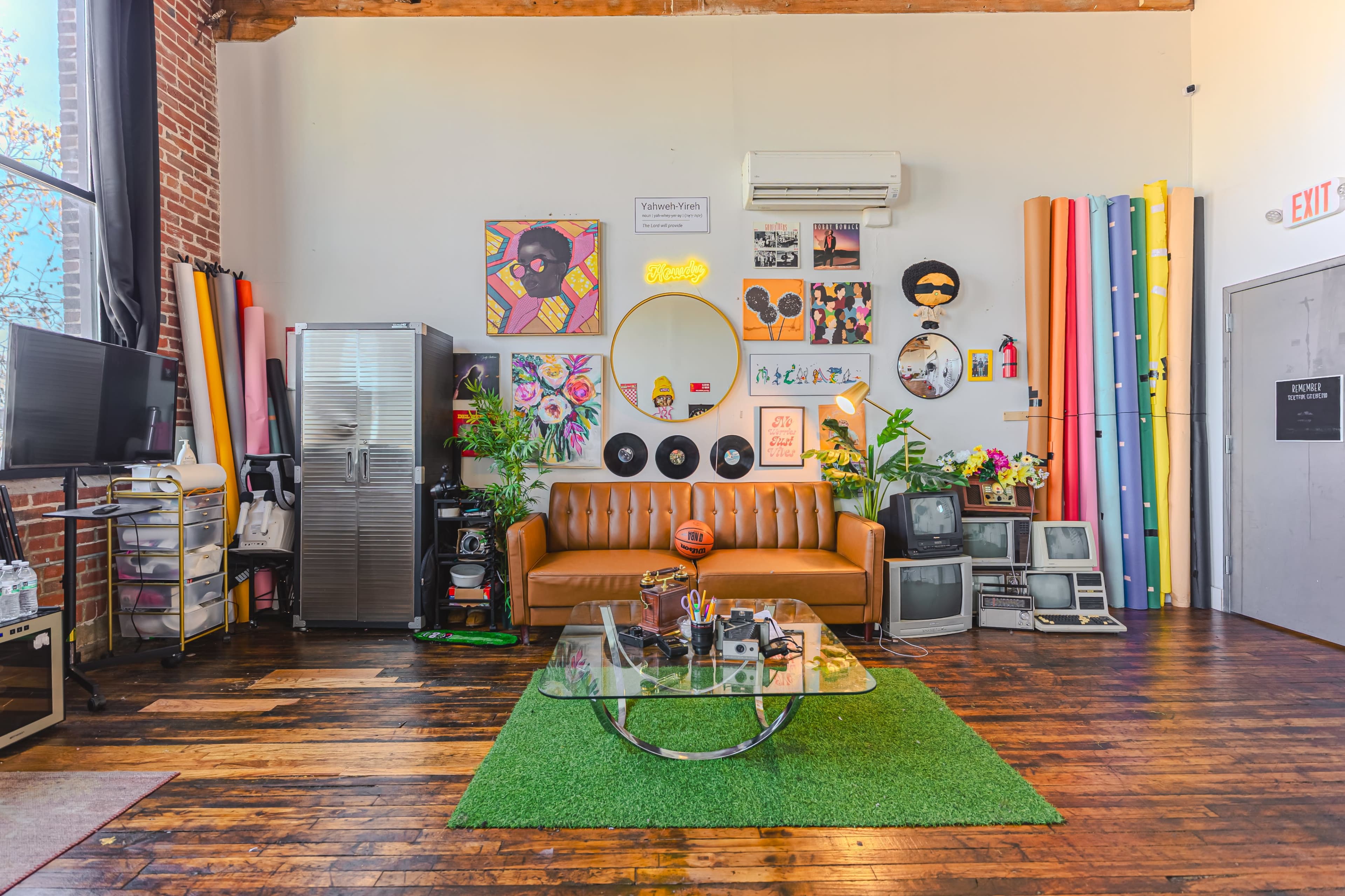 The image shows a vibrant urban studio space featuring a brown leather couch, colorful wall decorations, and various retro items arranged on a wooden floor.