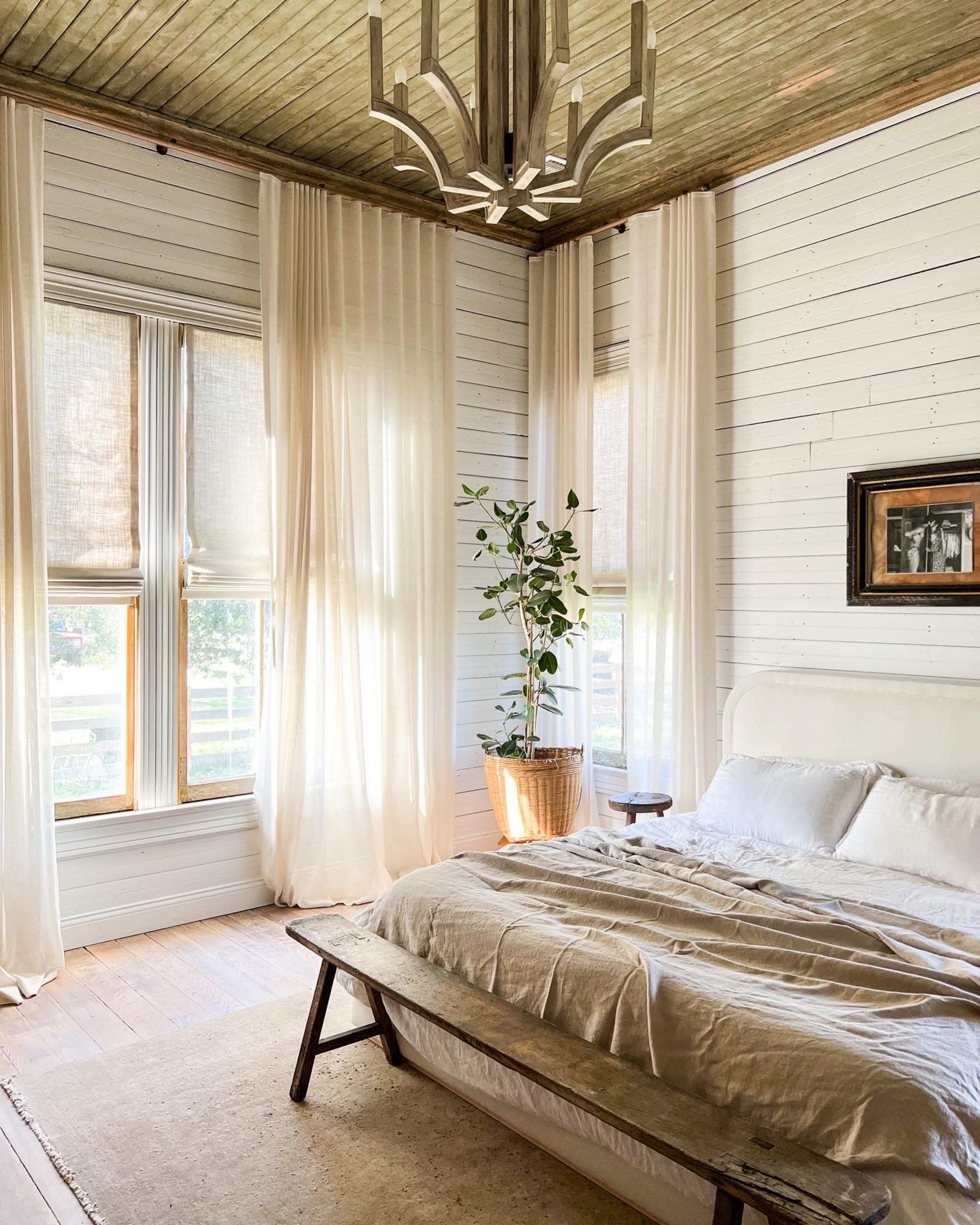A bright bedroom features a bed with a light linen cover, a wooden bench, and large windows draped with sheer curtains, accompanied by a potted plant and a framed picture on the wall.