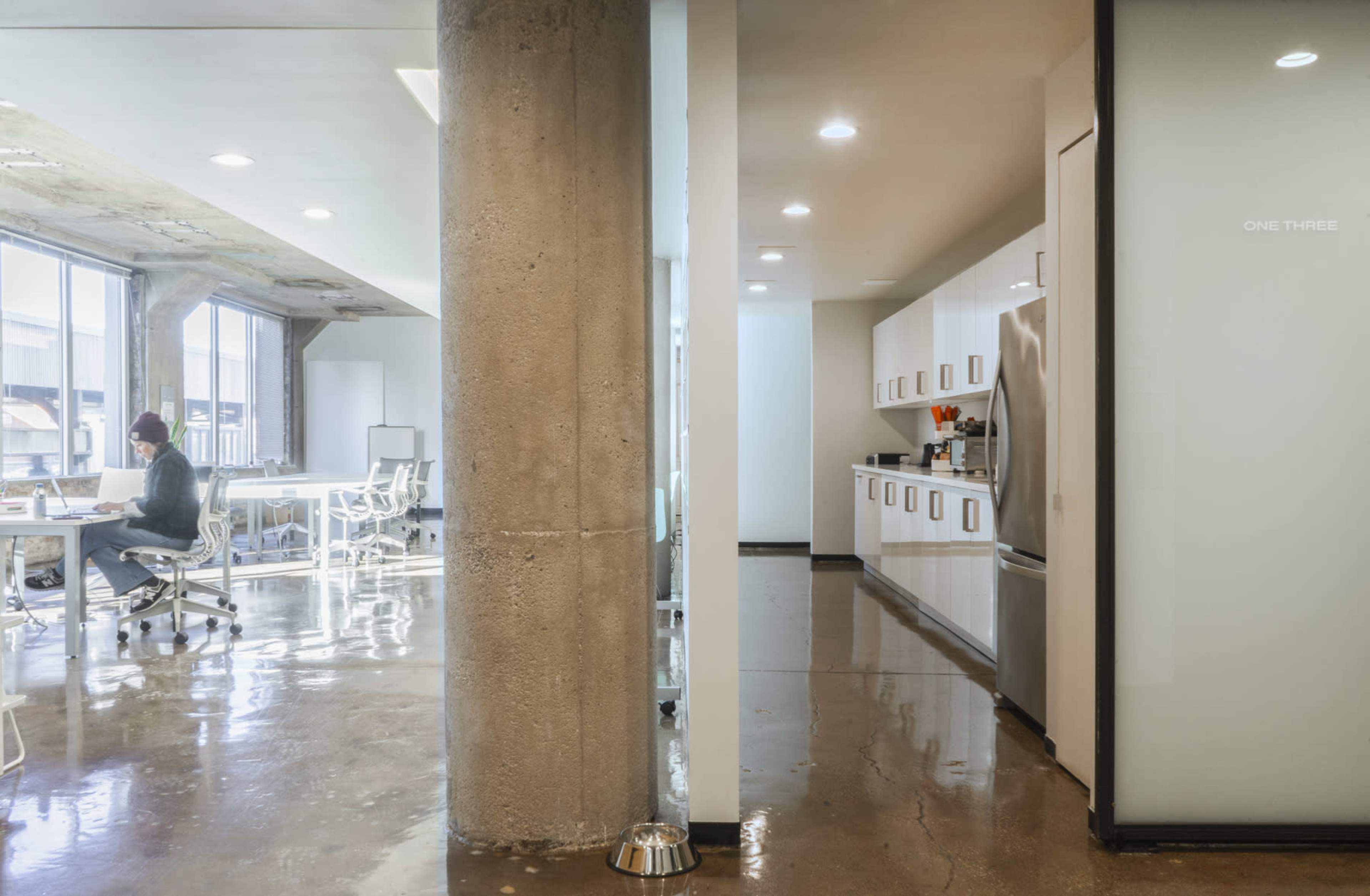 The image shows a modern office space divided by a concrete pillar, with a workspace featuring white desks and a separate kitchen area with white cabinets.