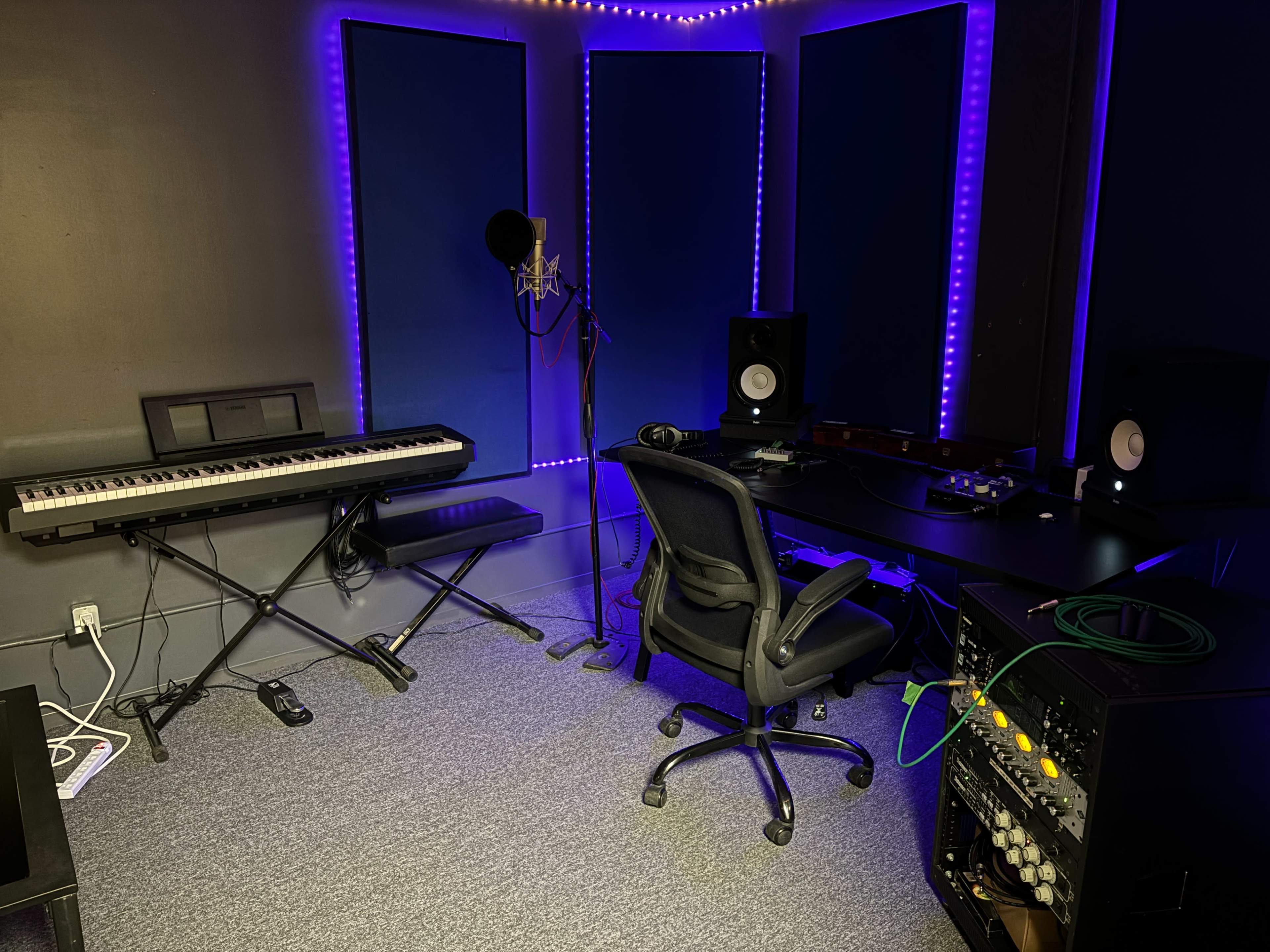 The image shows a small music studio featuring a keyboard, a microphone on a stand, studio monitors, and a mixing console, all illuminated by purple LED lights.