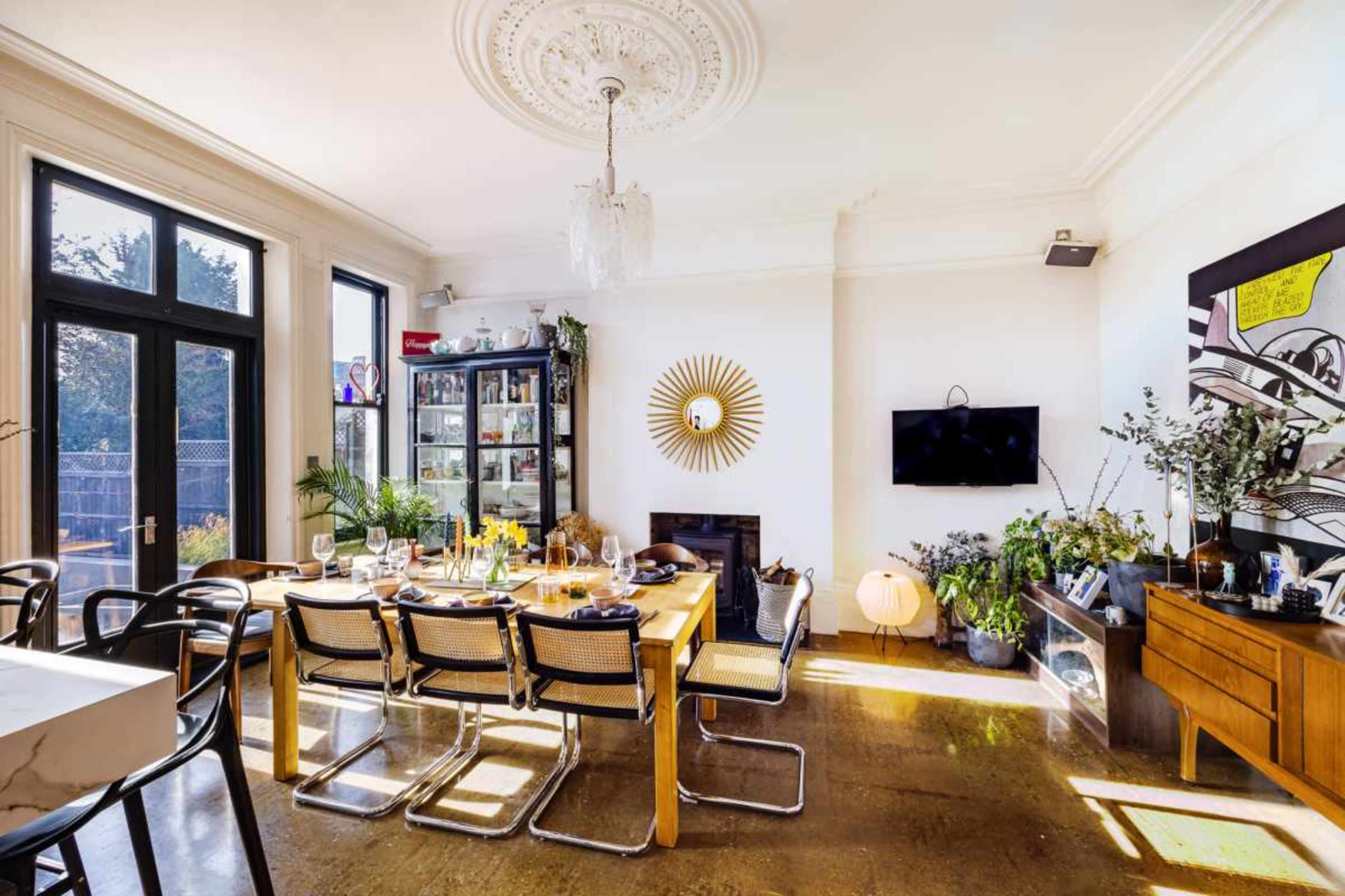 The image shows a well-lit dining room with a long wooden table, surrounded by modern chairs, and decorated with plants and wall art.