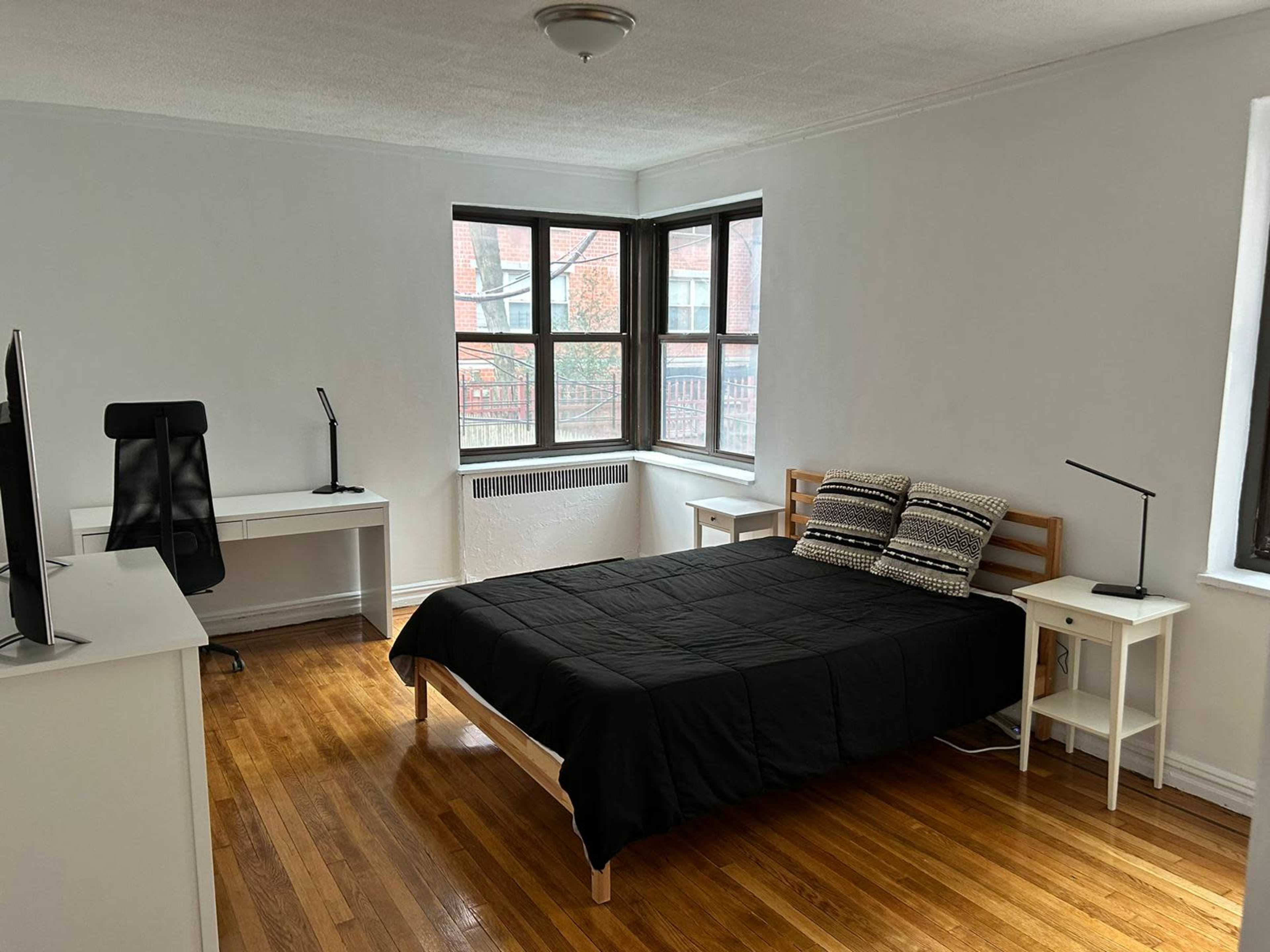 A bright bedroom with a wooden bed, a desk with a chair, and two lamps on white furniture against a backdrop of large windows.