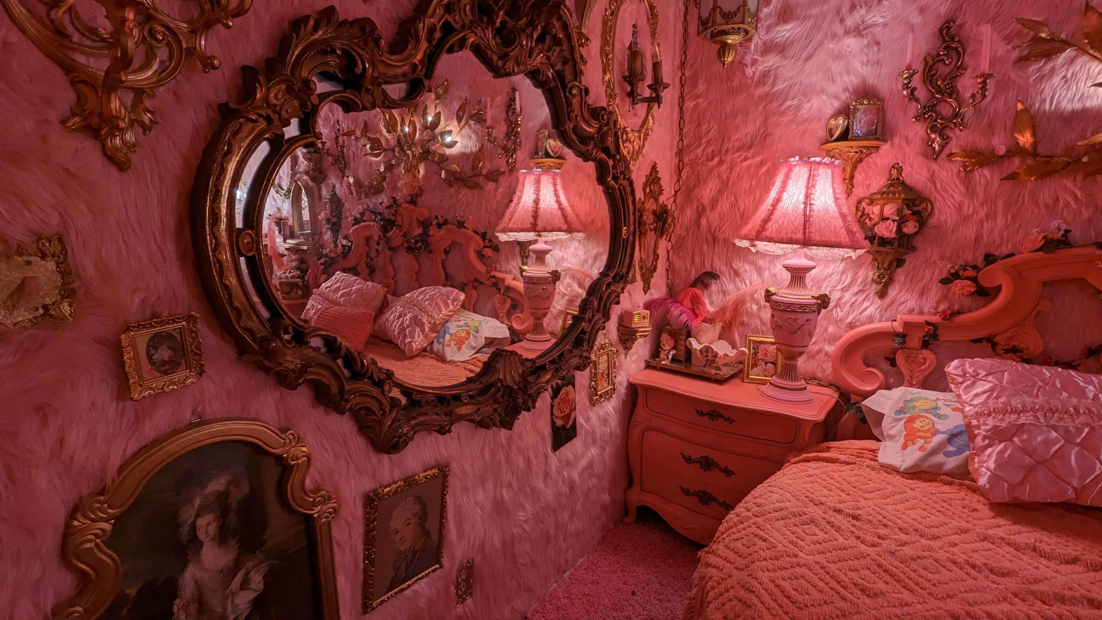 A pink-themed bedroom with a plush wall, ornate mirrors, and vintage-style decor, including a bed, bedside table, and framed artwork.
