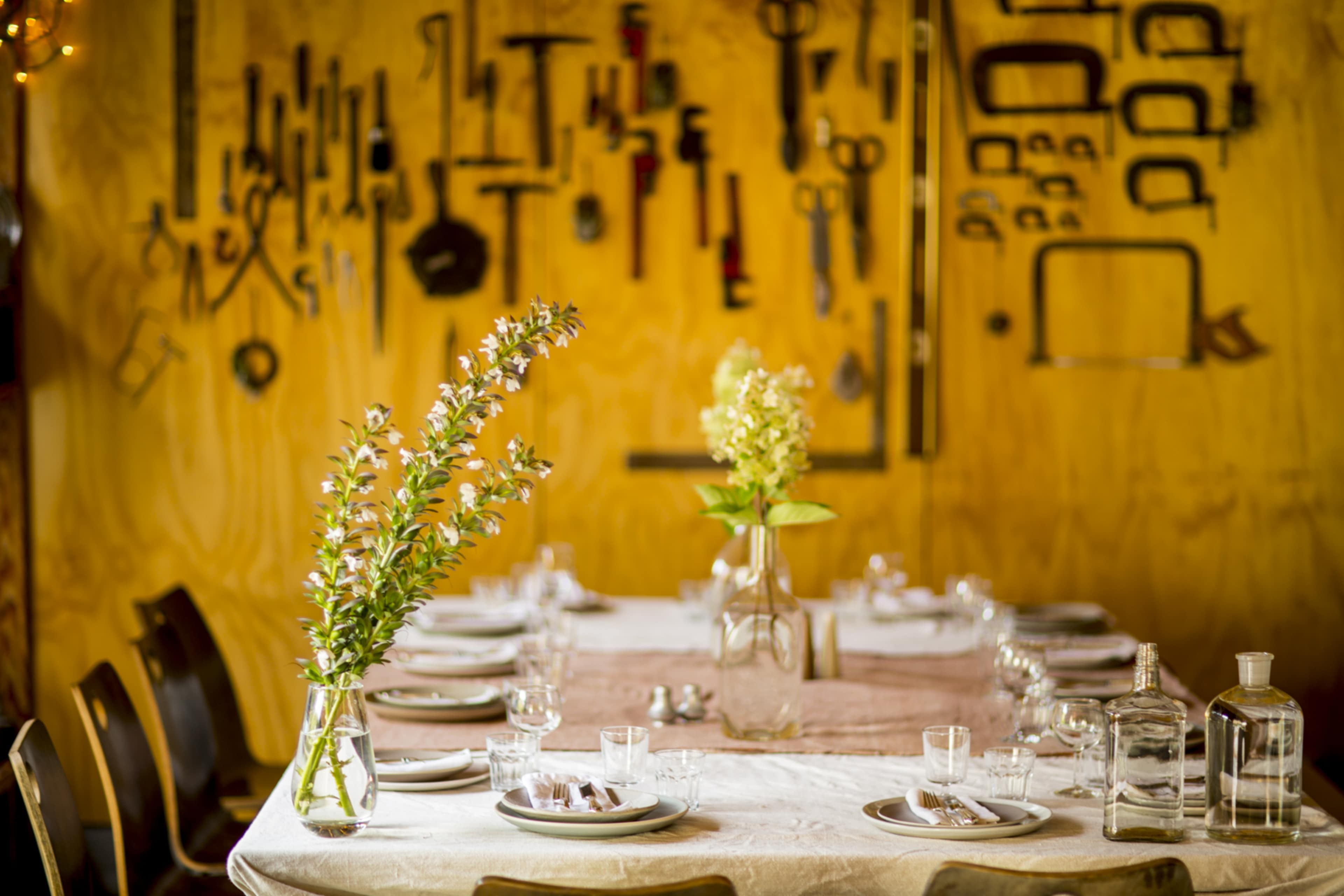 A dining table is set with plates and glasses, flanked by flower arrangements and surrounded by a wall displaying various tools.