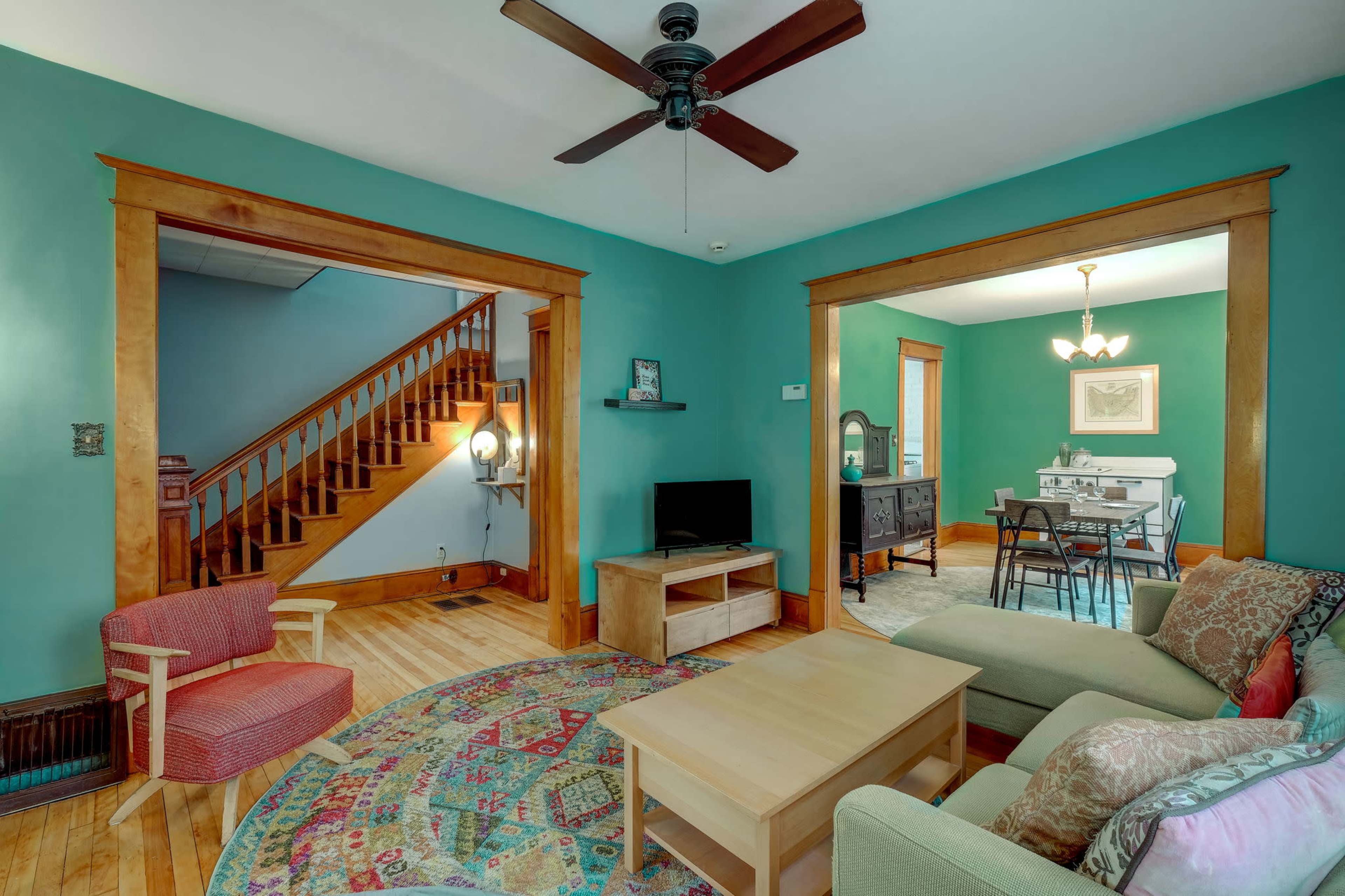 A living room features a teal wall, wooden trim, a ceiling fan, a coffee table, a couch, a chair, and a view of a staircase and dining area.
