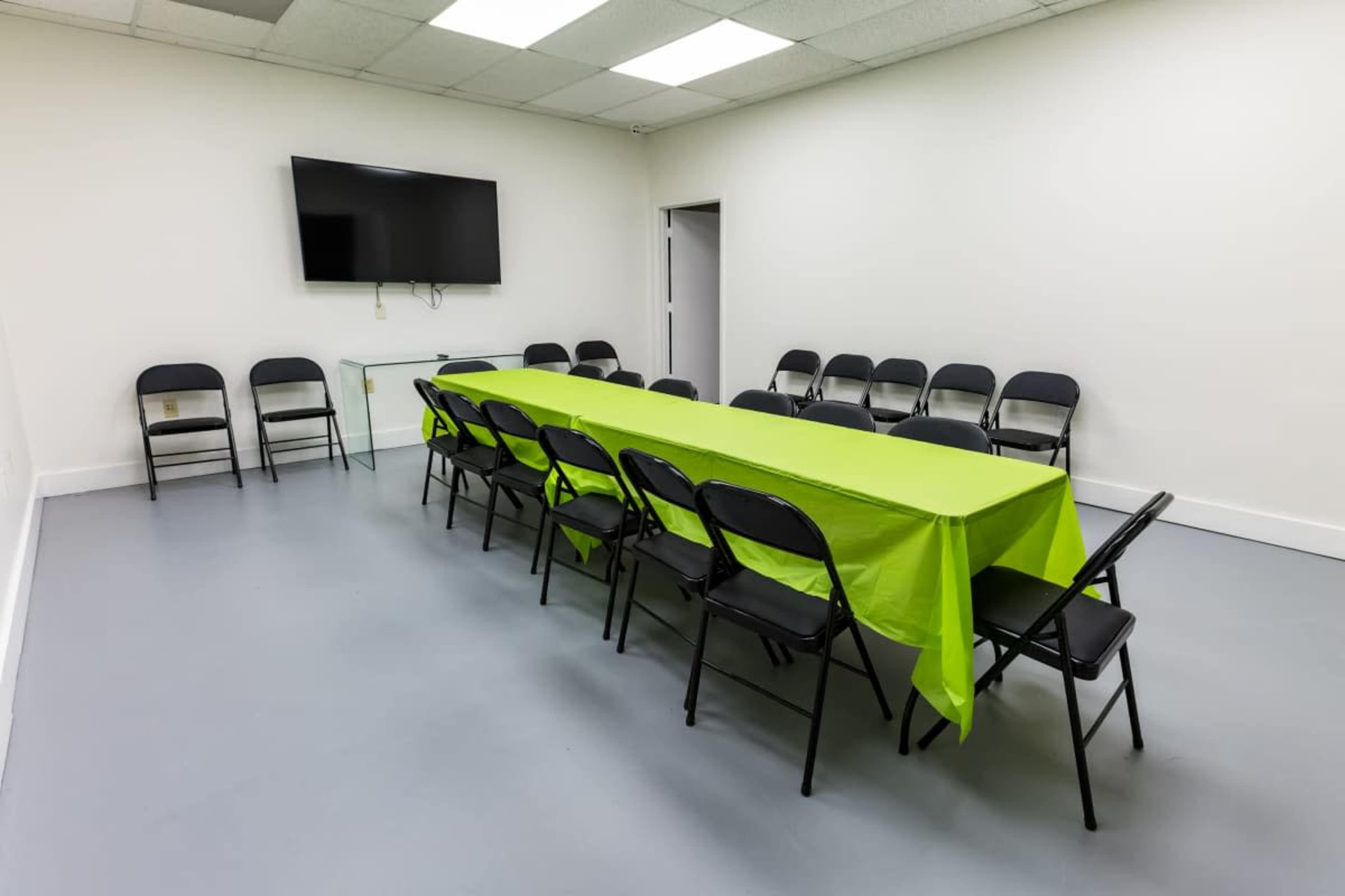 A rectangular table with a lime green tablecloth is set up in a white-walled meeting room surrounded by black folding chairs.