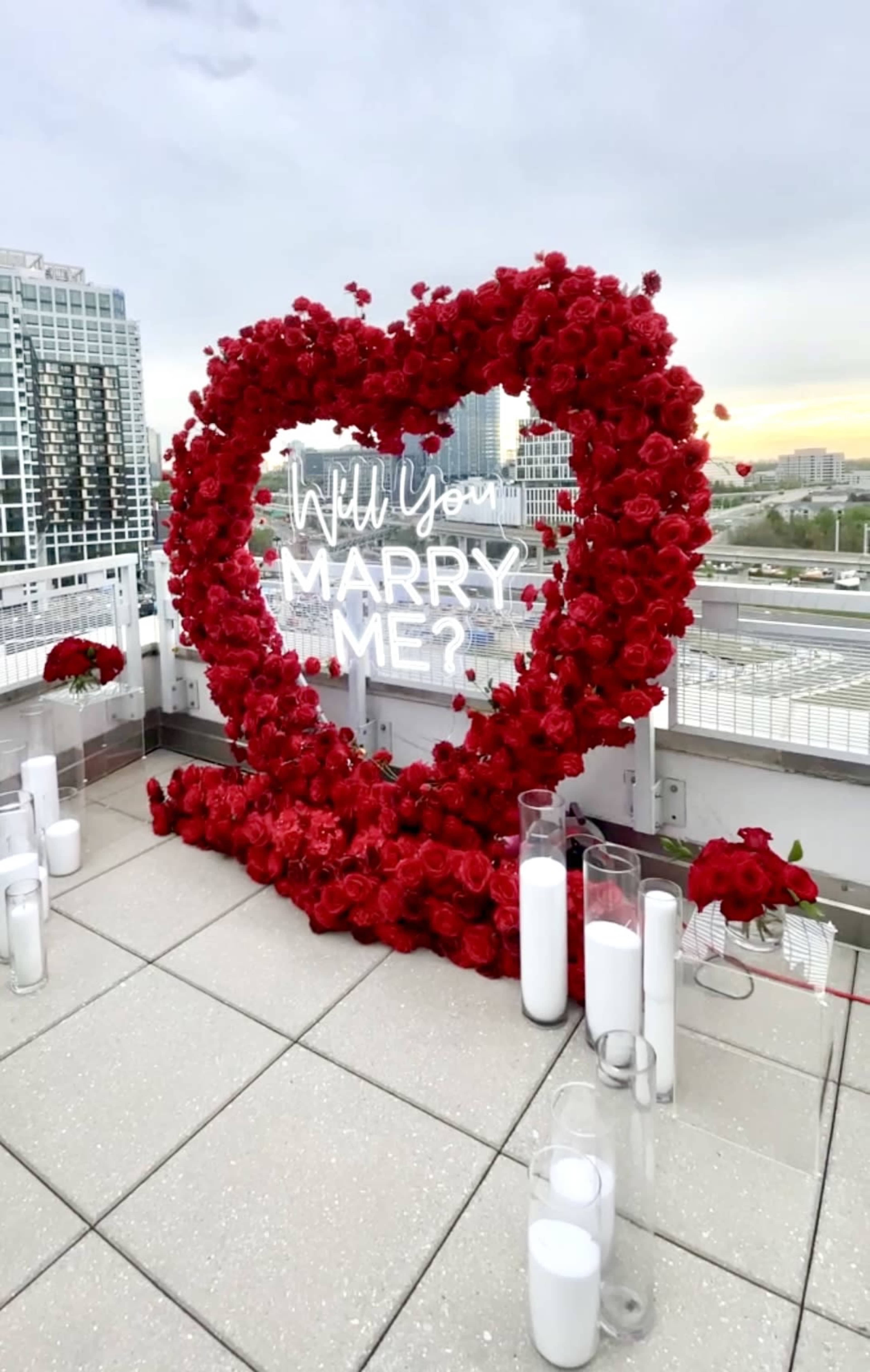 A heart-shaped arrangement of red roses surrounds a sign that reads "Will You Marry Me?" on a rooftop setting.