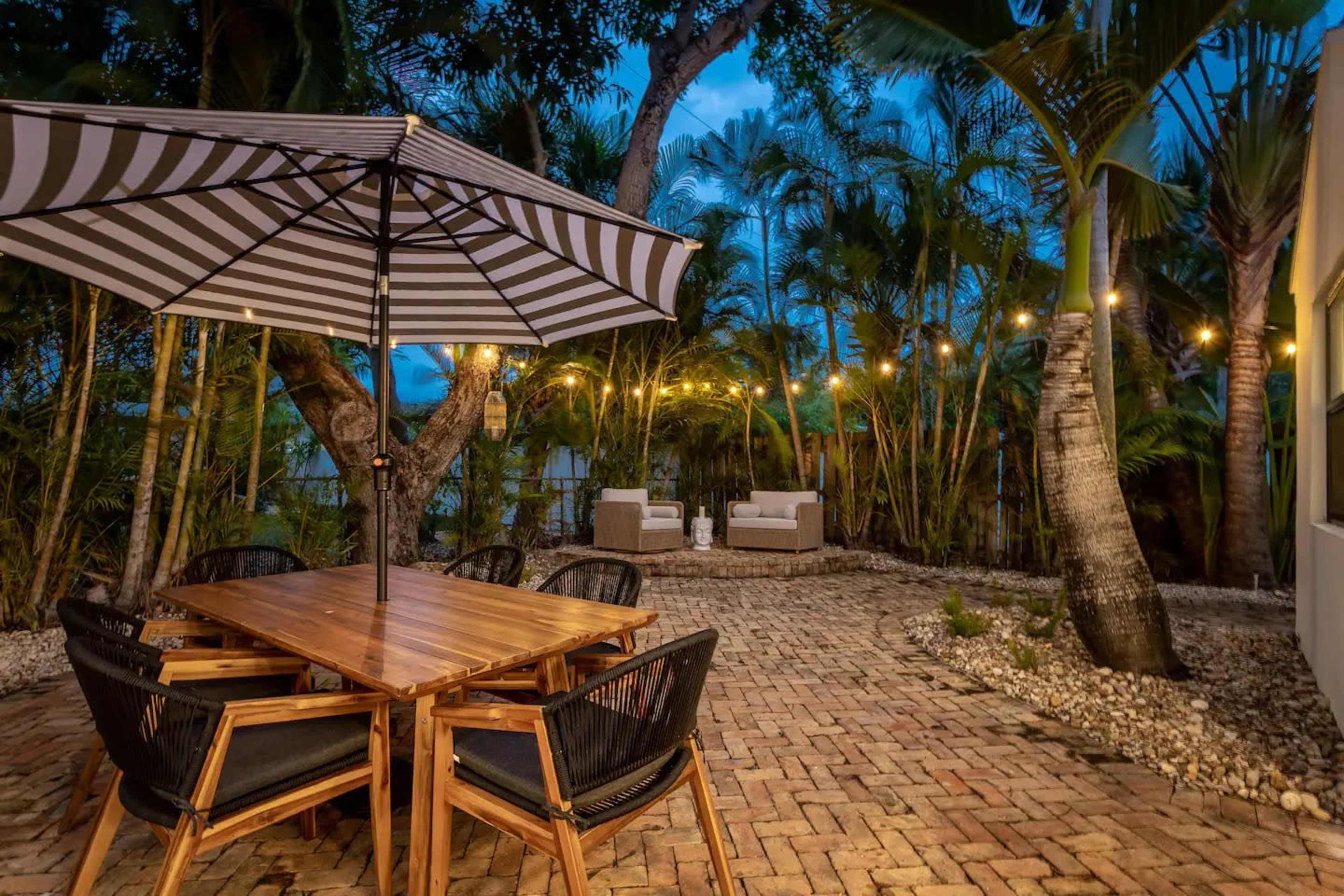 A wooden dining table with black chairs sits under a striped umbrella in a landscaped outdoor space, adorned with string lights and surrounded by palm trees.