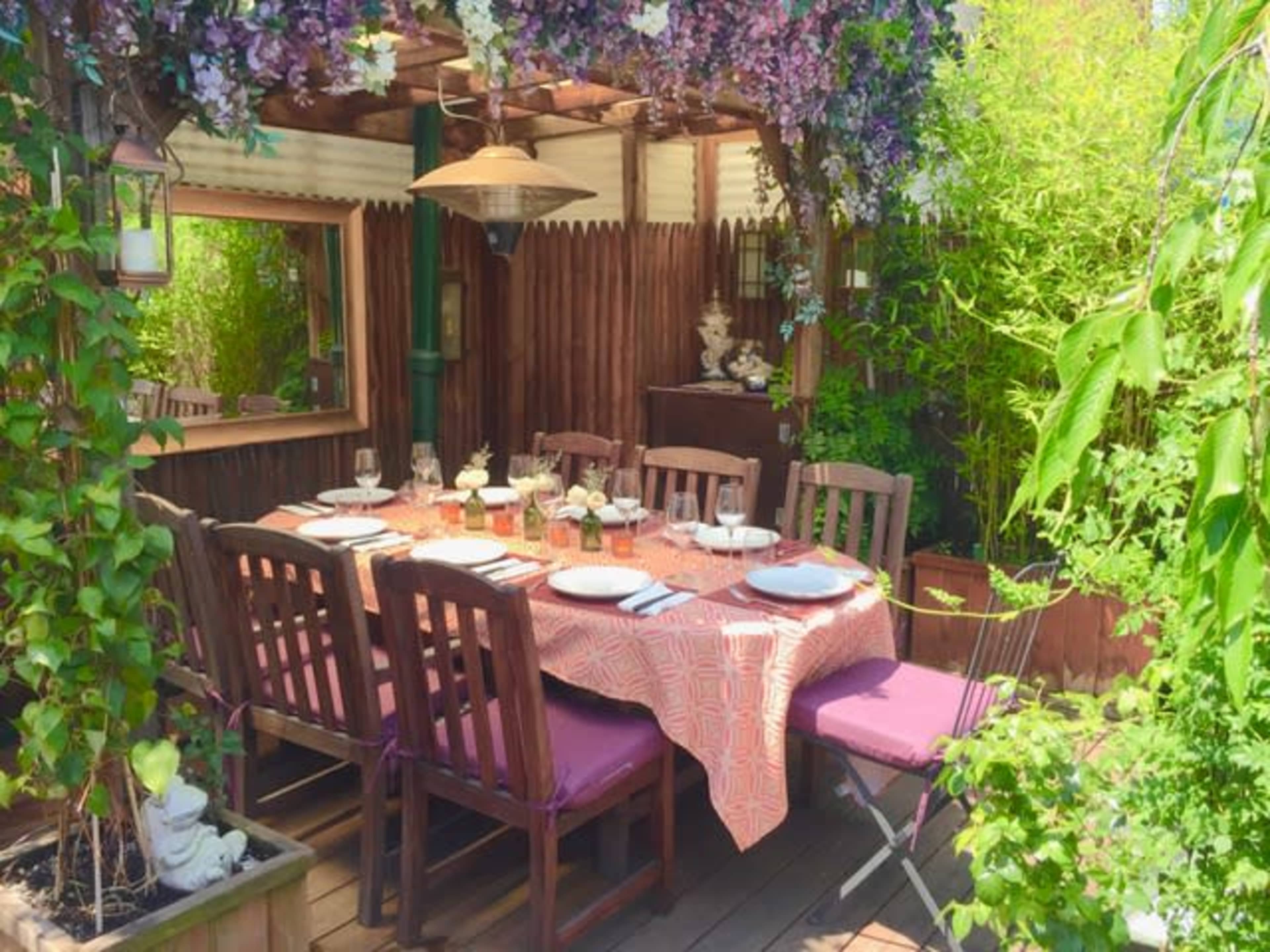 A dining table is set with plates and glasses in a garden gazebo surrounded by greenery and flowers.
