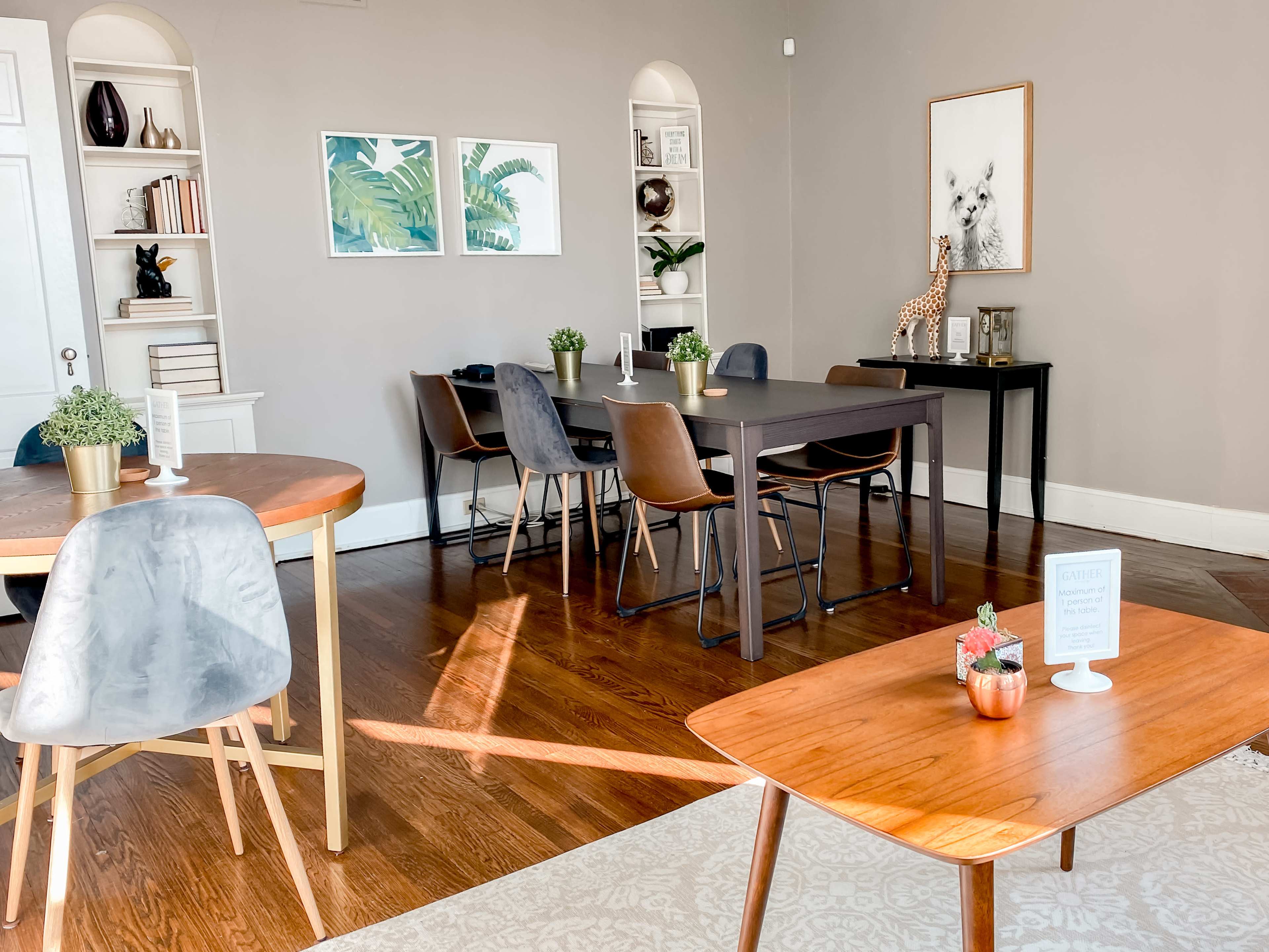 The image shows a well-lit room with a mix of wooden furniture, including tables and chairs, along with decorative plants and framed artwork on the walls.