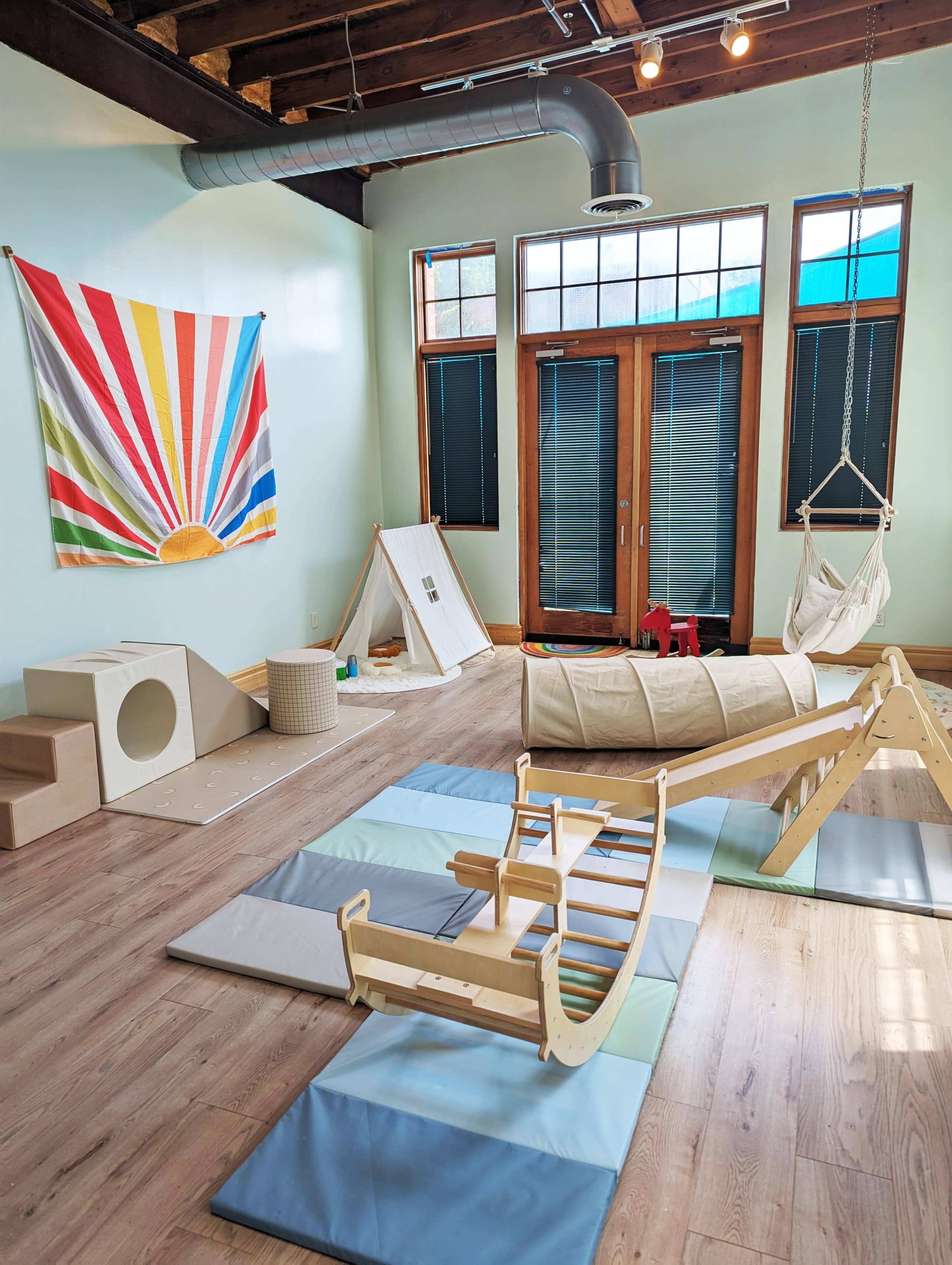 The room features soft play equipment, a colorful wall mural, and large windows with black-out shades.