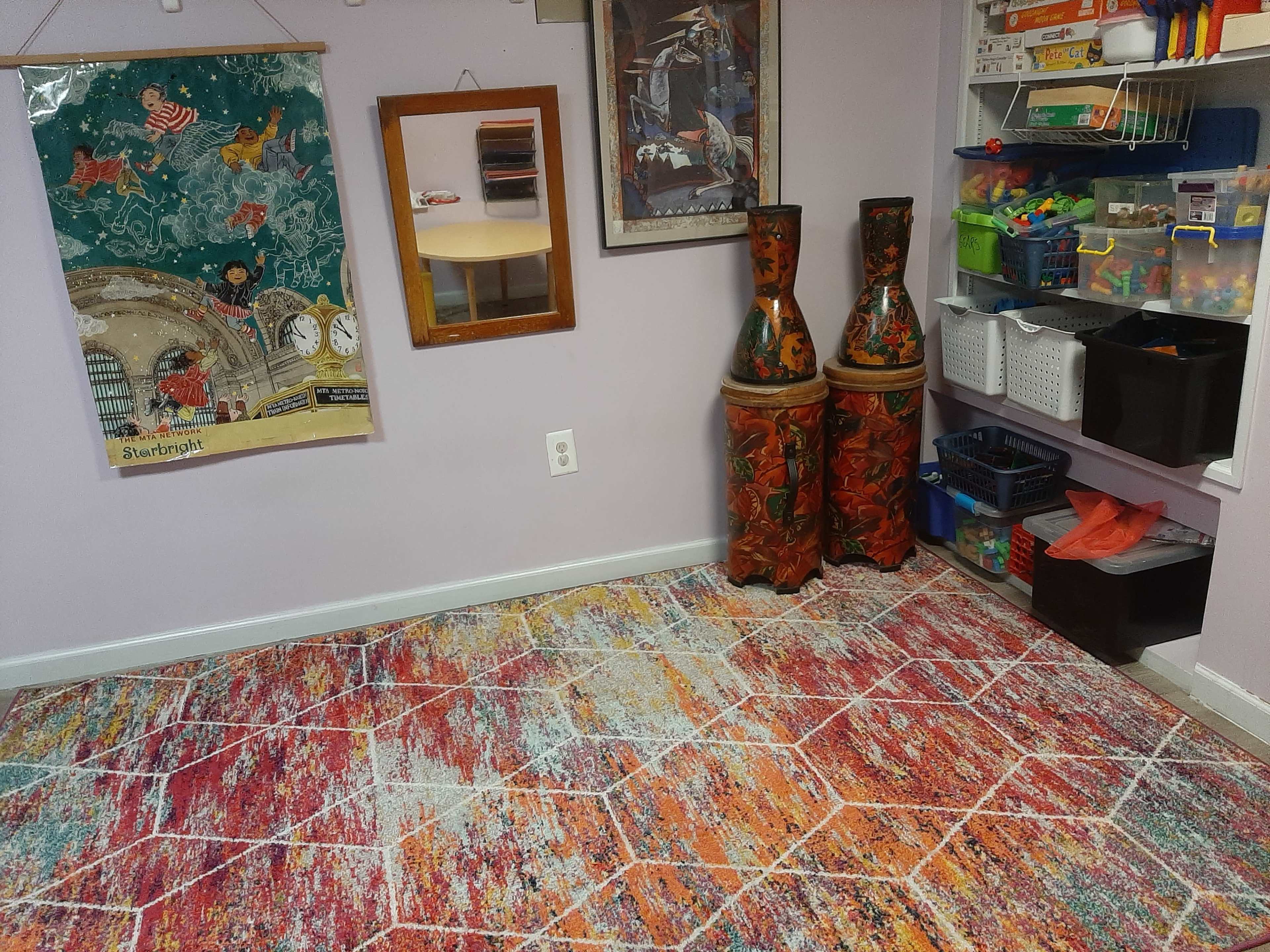 A brightly colored room with a patterned rug, a mirror on the wall, and two decorative vases beside a storage unit filled with bins.