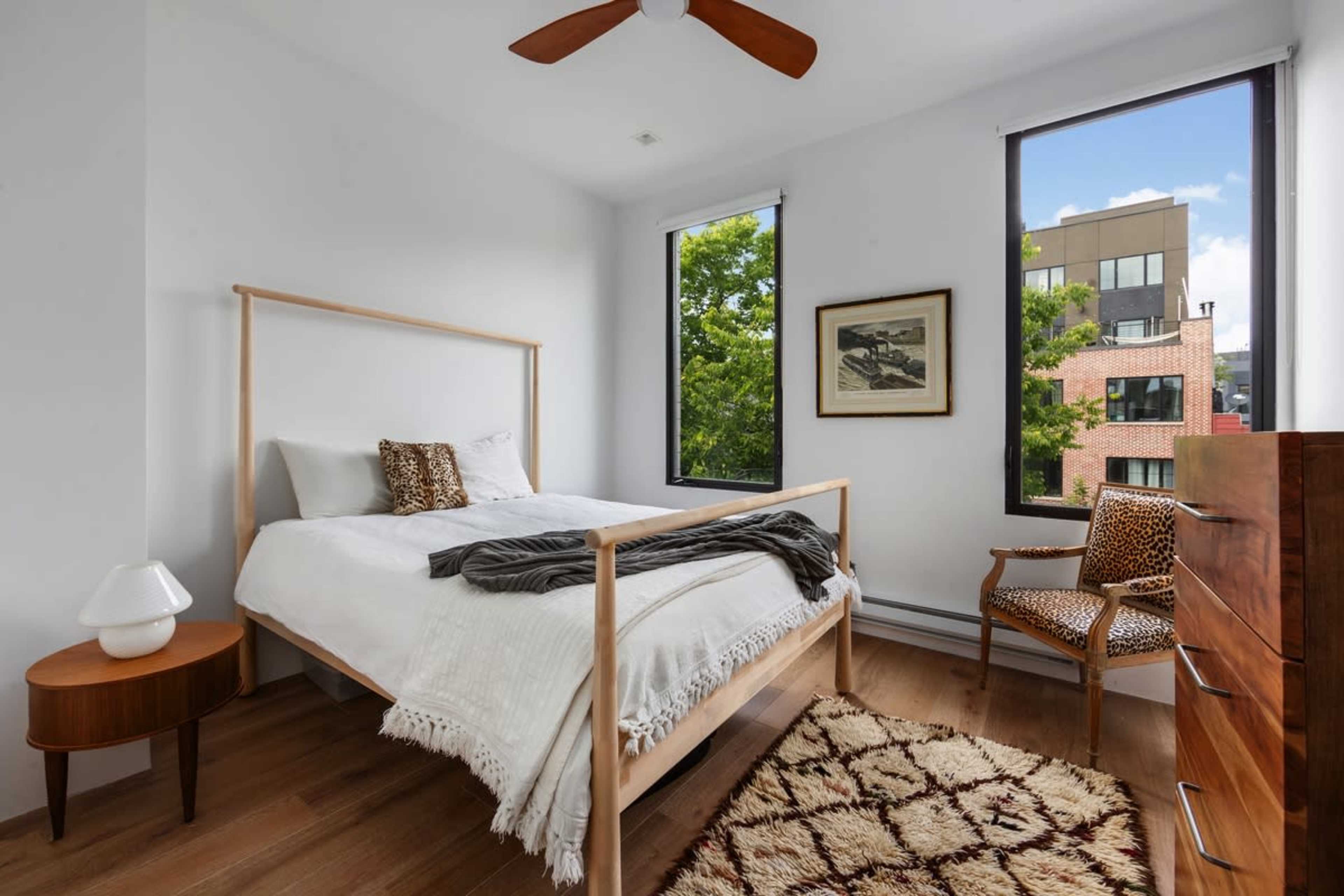 A modern bedroom features a wooden bed with white bedding, a side table with a lamp, a patterned chair, and large windows overlooking a cityscape.