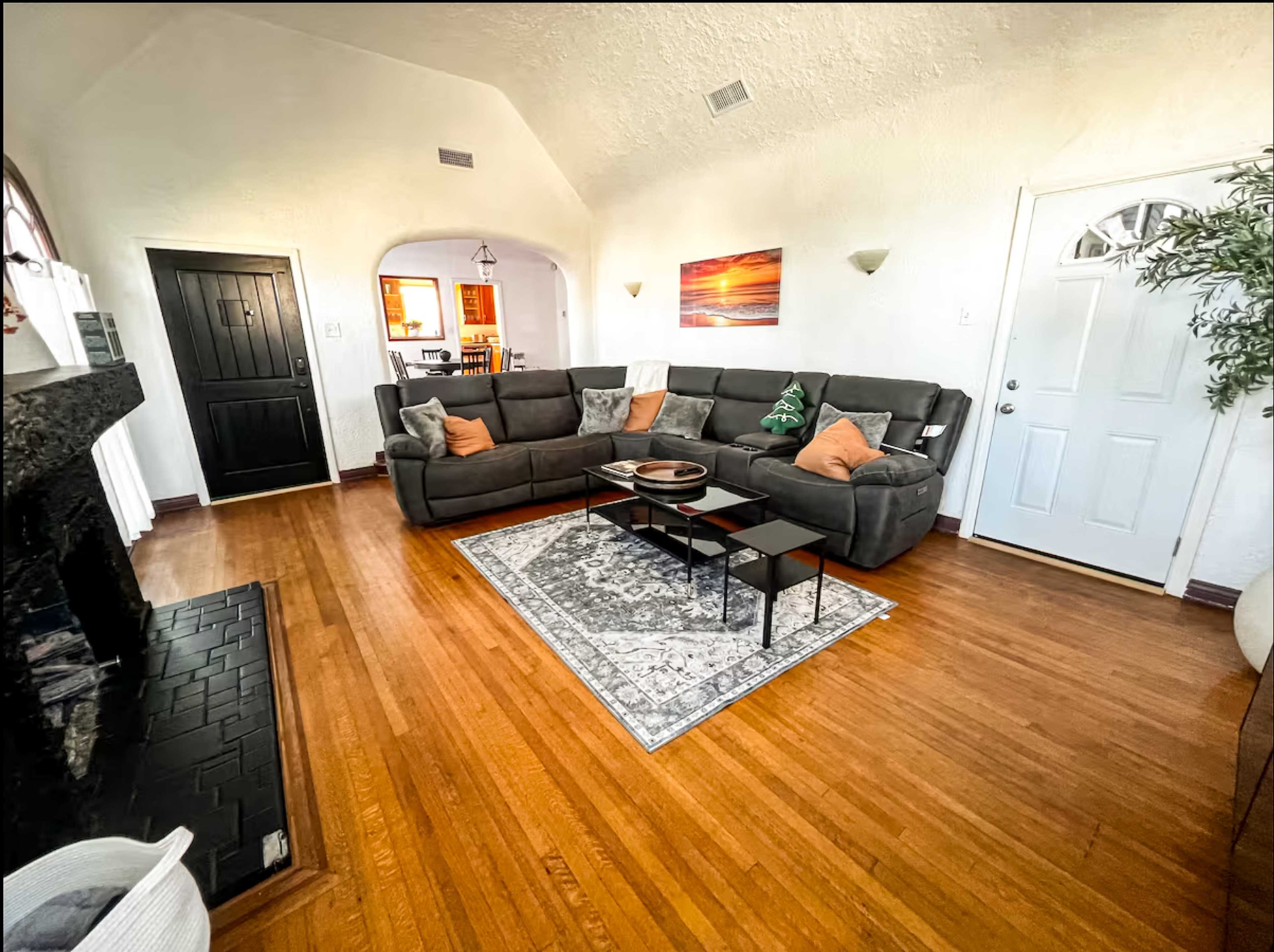 The image shows a living room with a gray sectional sofa, a black coffee table, a decorative rug, and a fireplace, all set against white walls and hardwood floors.