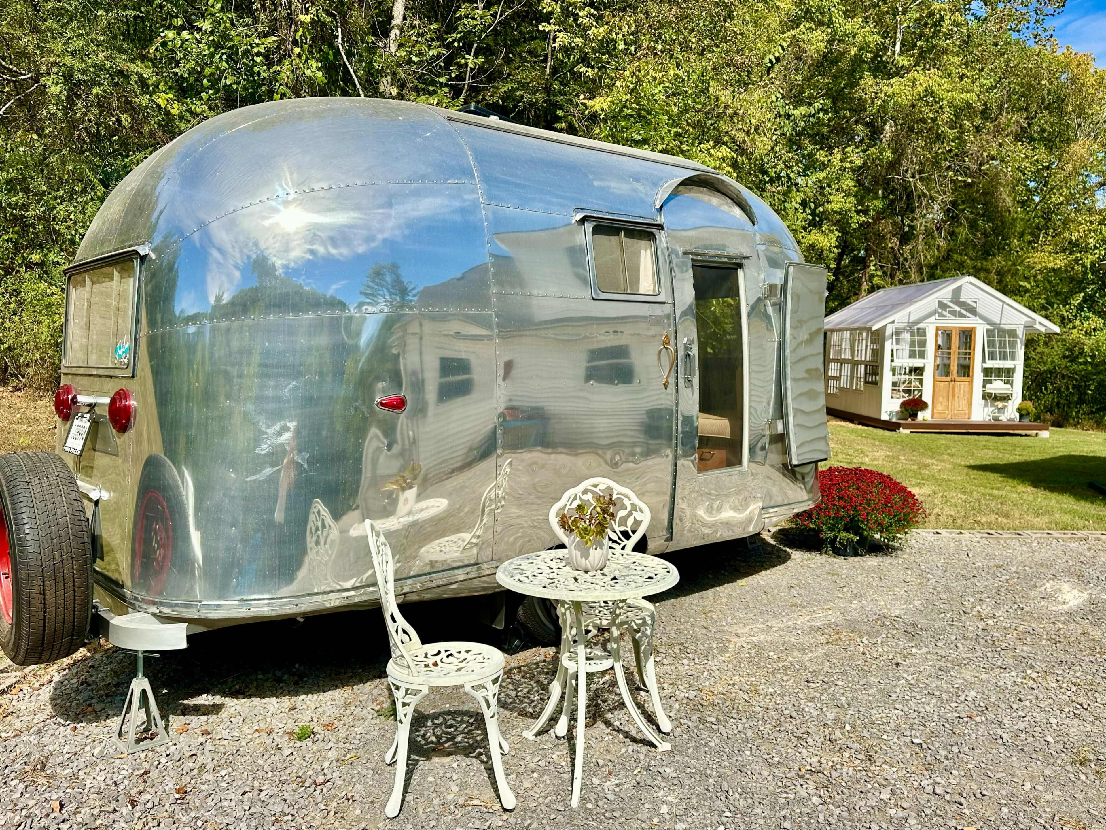 A shiny vintage Airstream trailer is parked on a gravel driveway, next to a small white building surrounded by greenery and flowers.