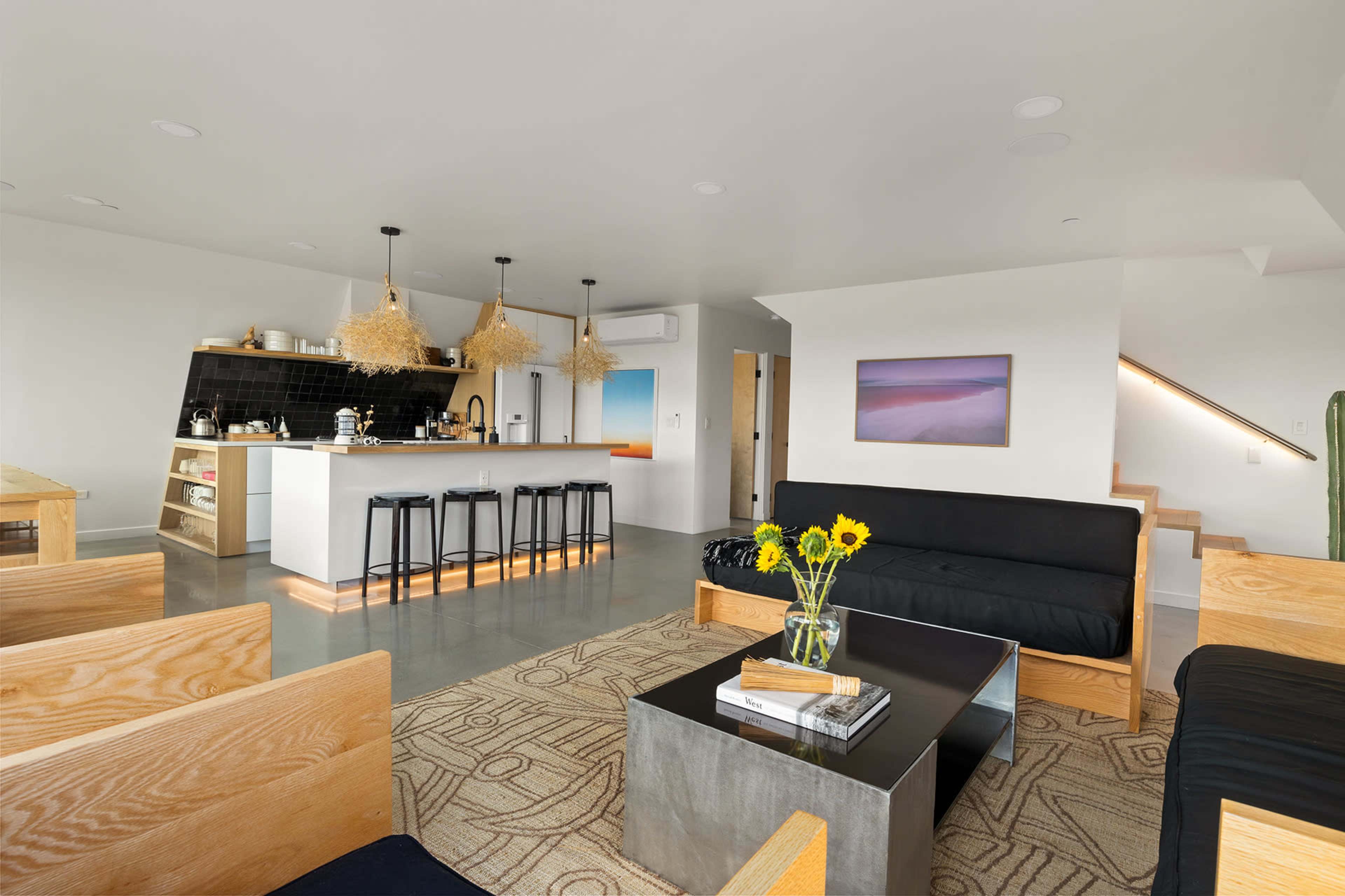 The image shows a modern open-concept living space with a kitchen, dining area, and seating area featuring minimalist furniture and decorative plants.