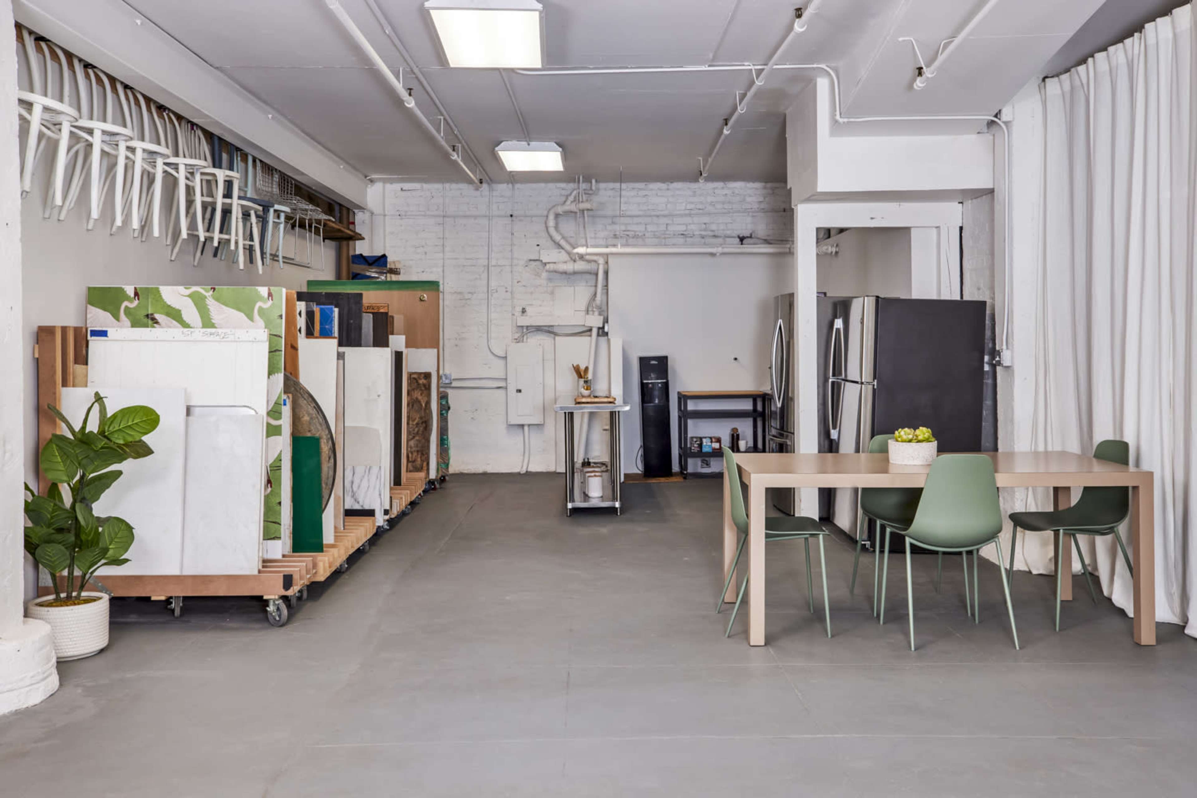 A spacious room features several refrigerators on carts along one side, a table with chairs in the center, and a partitioned area with a curtain.