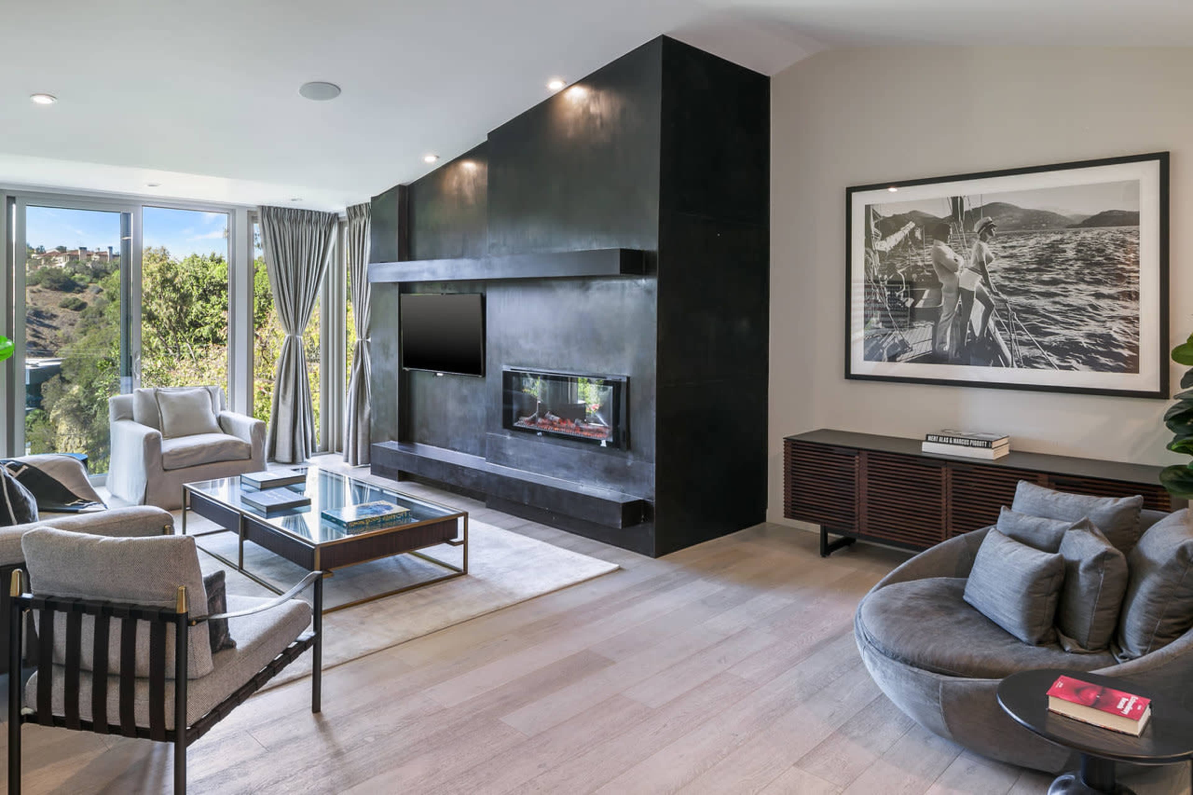 The living room features a modern design with a black fireplace, large windows overlooking greenery, and a mix of comfortable seating.