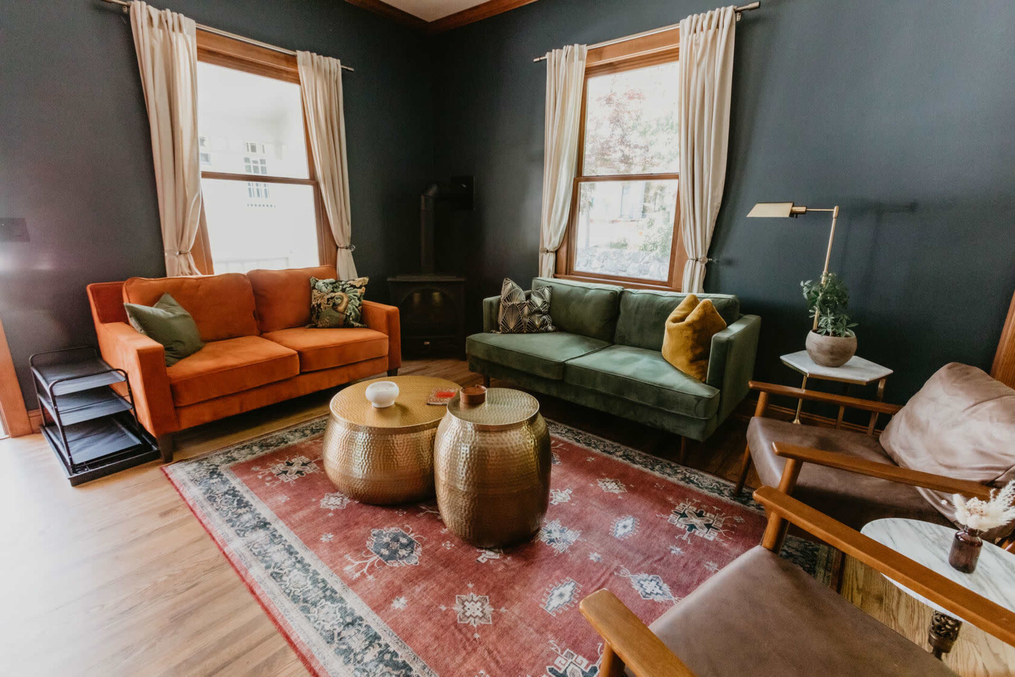 The living room features two sofas in orange and green, a patterned rug, and two round metal coffee tables against a dark wall.