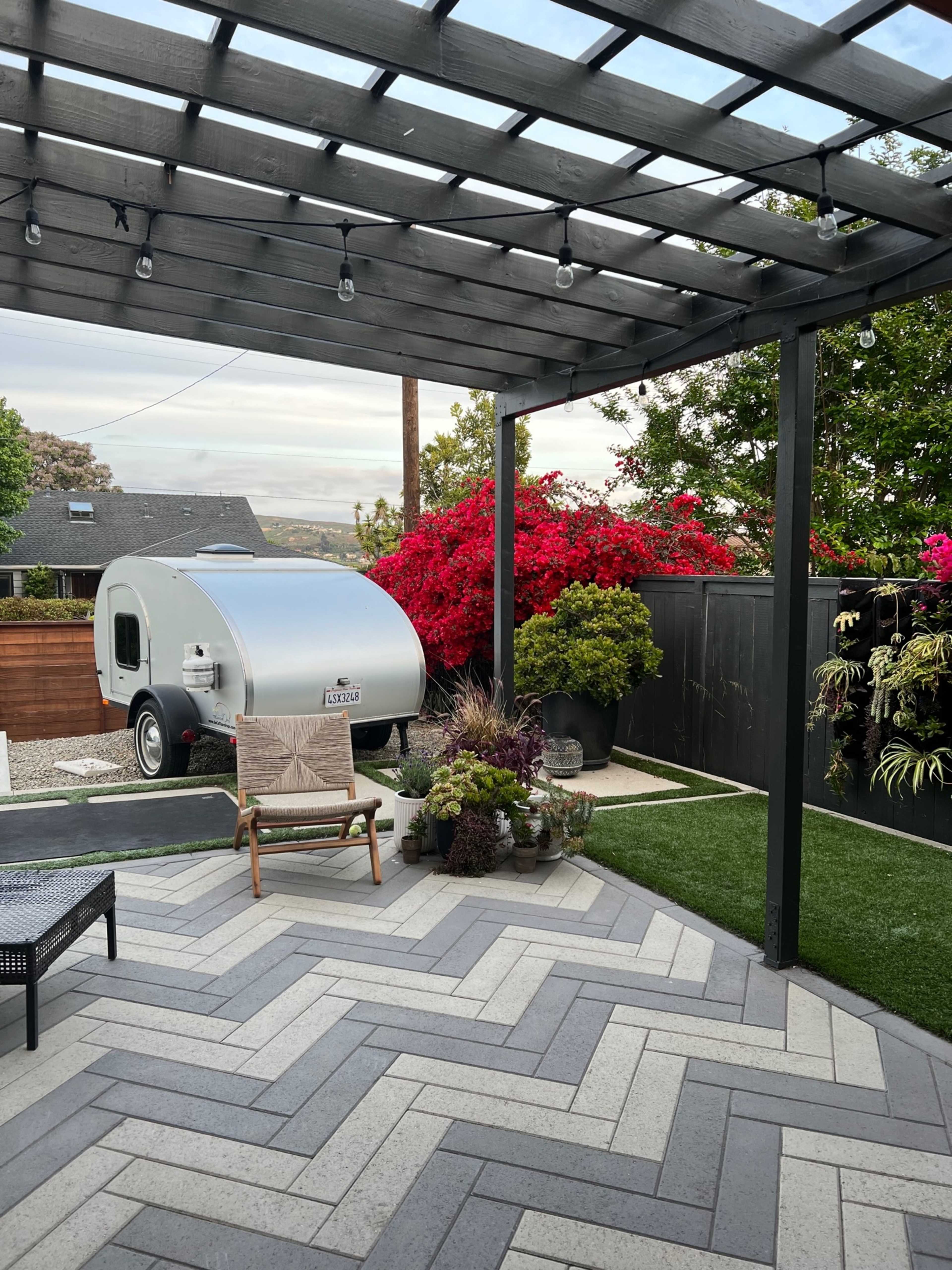 A small silver trailer is parked under a wooden pergola surrounded by colorful flowers and a well-maintained patio with herringbone-patterned tiles.