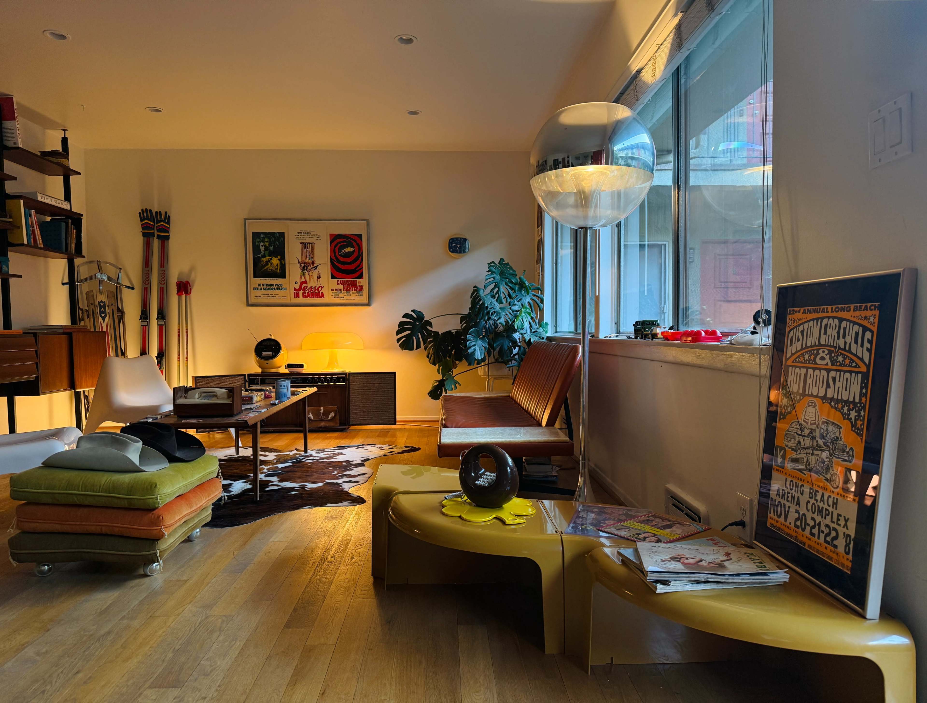 A mid-century modern living room features a combination of wood furniture, a colorful rug, and framed posters on the walls, with large windows allowing natural light to fill the space.