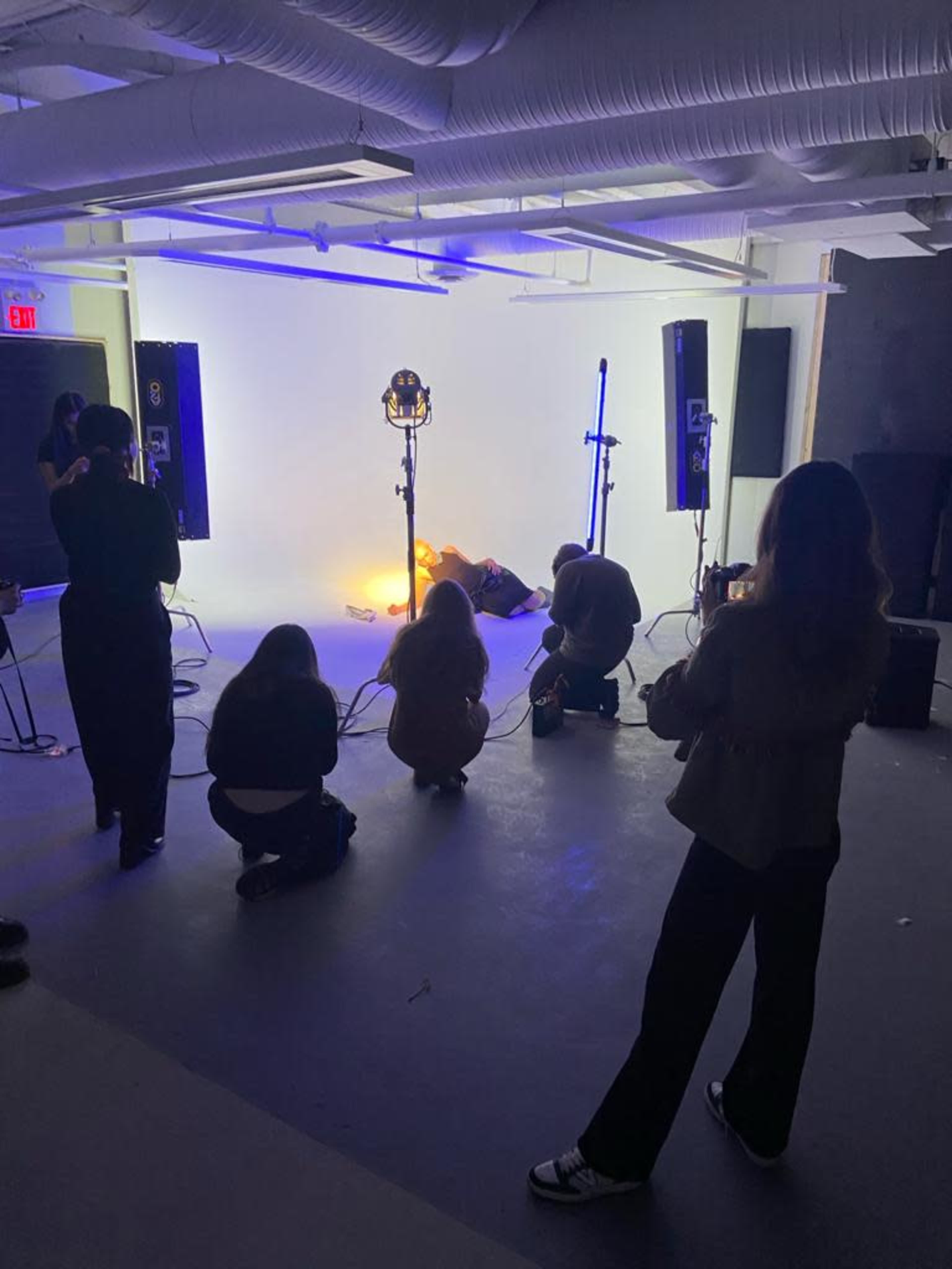 A group of people is arranged in a studio, capturing a scene with a model lying on the floor amidst lighting equipment and a plain backdrop.