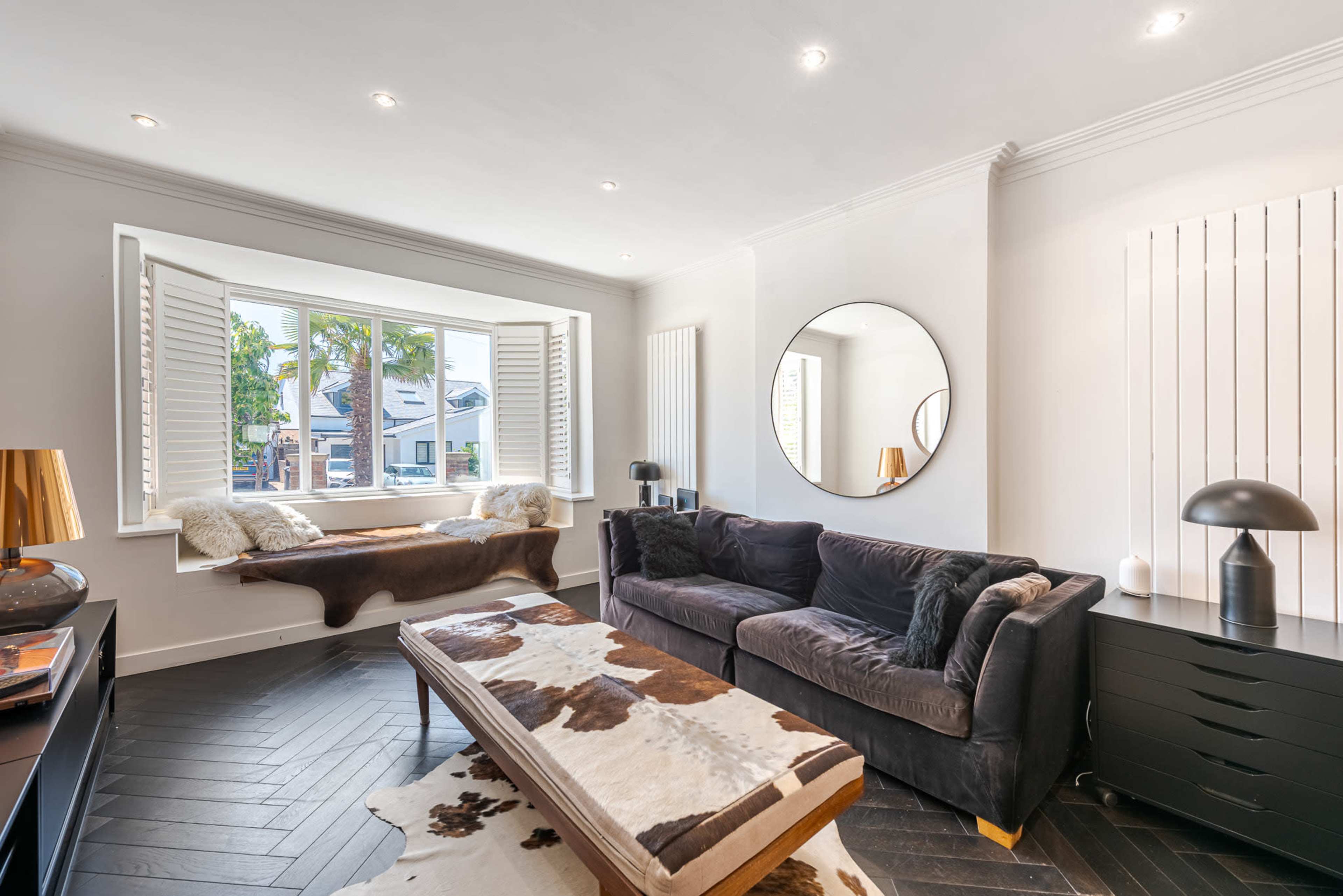 The image features a modern living room with a large window seat, a dark sectional sofa, a round mirror, and a coffee table on a patterned rug.