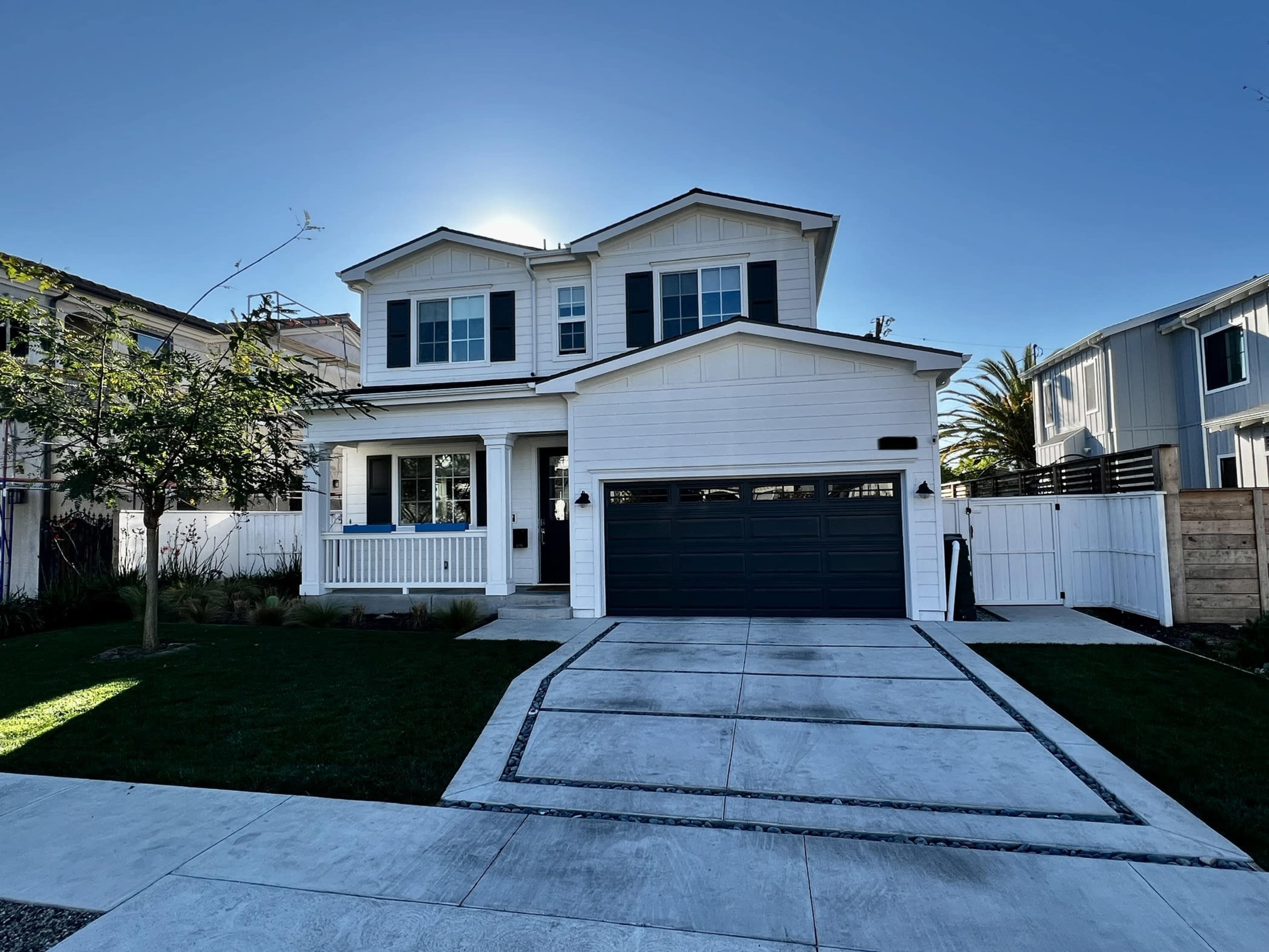 A two-story white house with a dark gray garage door and blue window shutters stands in a landscaped yard under a clear sky.